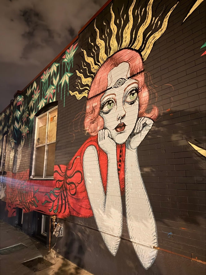 A large street art mural painted on a dark brick building wall in downtown denver at night, featuring a stylized woman with pink hair, white skin, a red top, and gold flame-like shapes behind her head. Photo taken by Brooke Gregson.