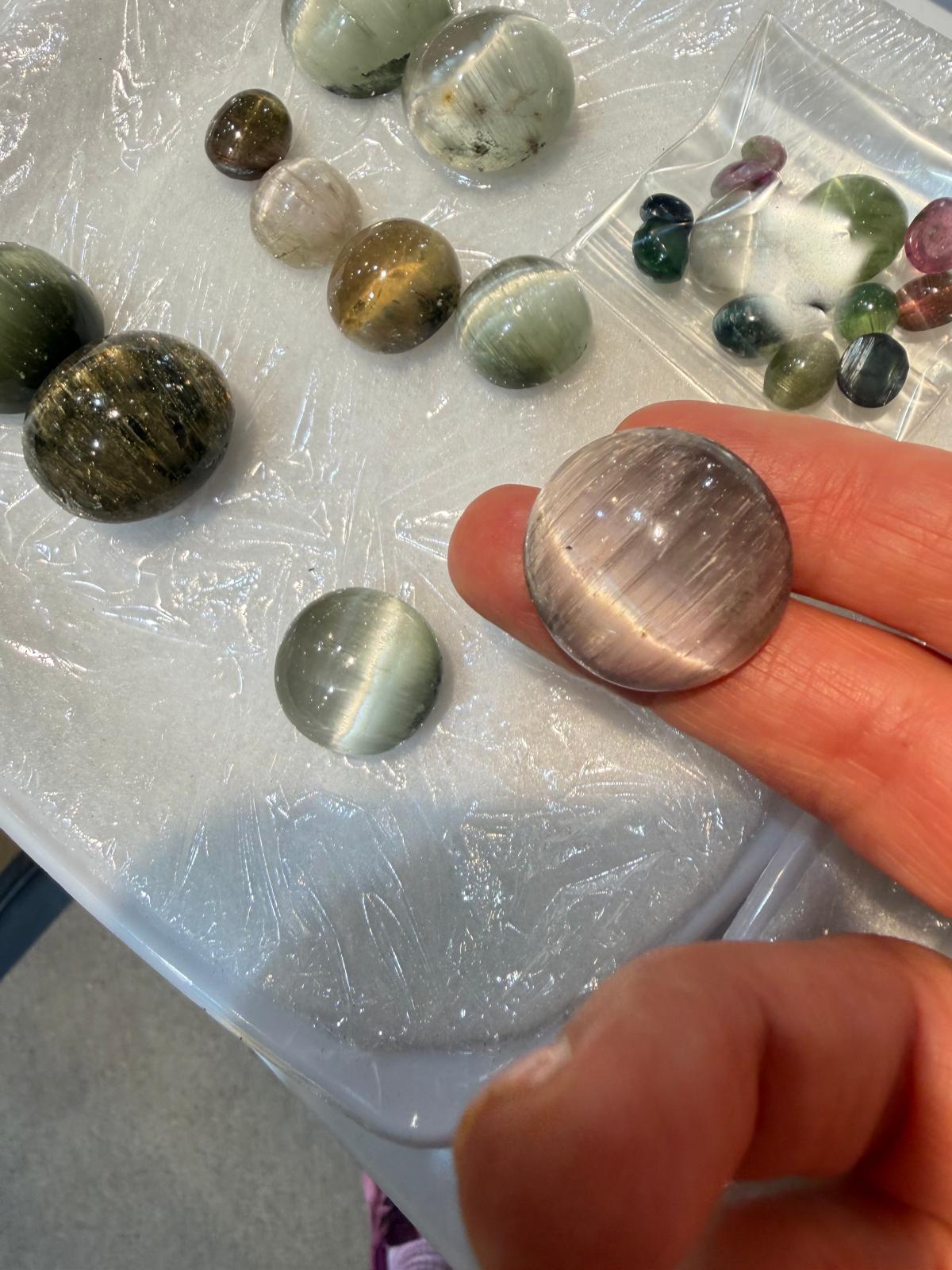 A hand holding a polished cat's eye gemstone, with several other polished stones and a small bag of loose stones arranged on a clear plastic-covered surface.