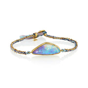 A gold-plated bracelet featuring a large, teardrop-shaped iridescent boulder opal stone with blue, purple, and green flashes, set on a braided blue and gold cord with a small tassel.