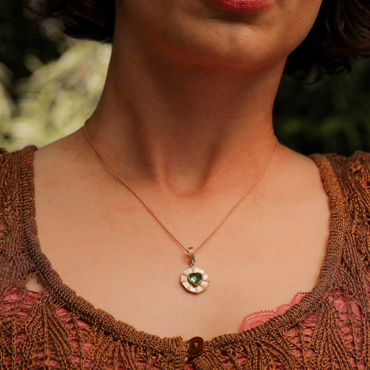 A close-up lifestyle shot of a woman wearing a delicate gold chain necklace with a heart-shaped pendant featuring a green heart in the center and textured hand engraved gold around it.