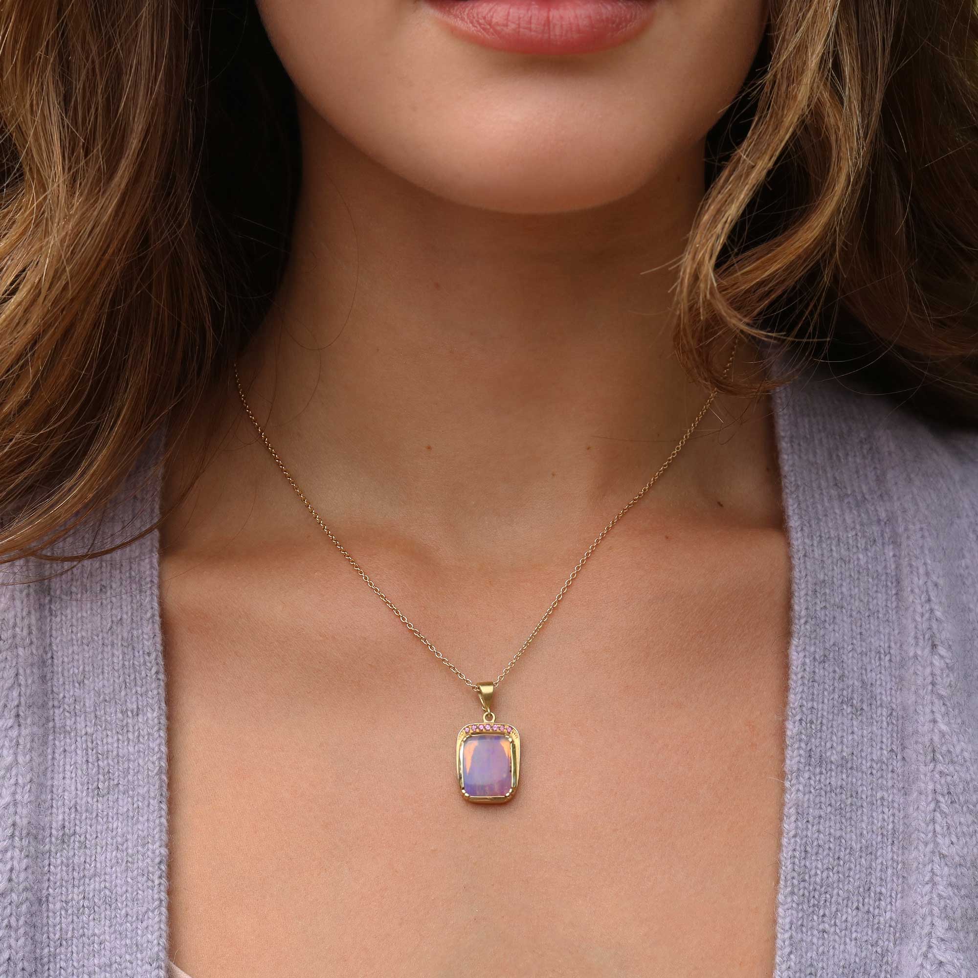 A close-up of a woman wearing a delicate gold chain necklace with a rectangular iridescent fossilised wood opal pendant that shifts between lavender and purple, set against her collarbone and a light purple cardigan.