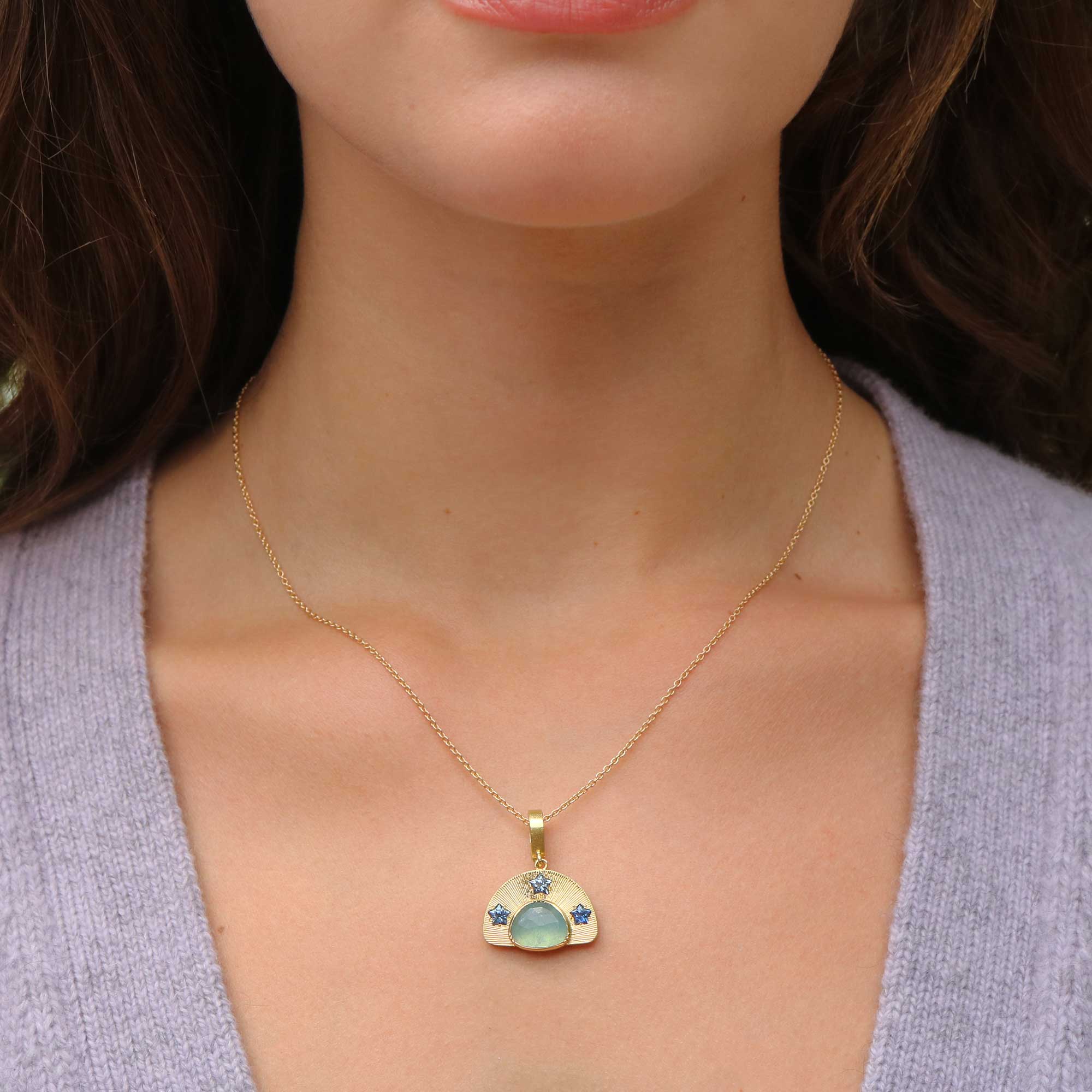 A close-up of a model wearing a delicate gold necklace with a small pendant featuring a light blue-green aquamarine stone and star shaped sapphire blue stones, paired with a light purple knit top.