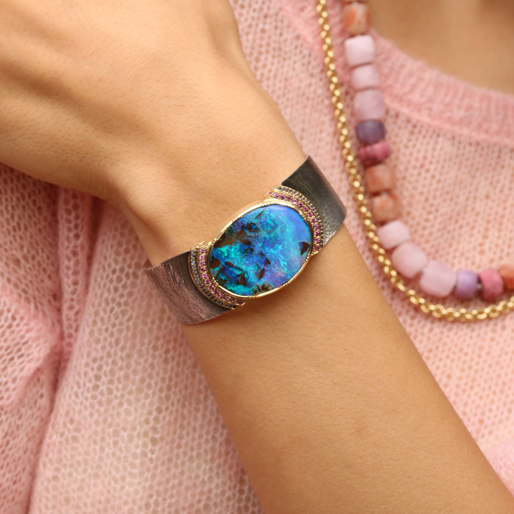 A close-up of a person's wrist wearing a wrap-style cuff bracelet with a large oval iridescent blue-green opal center and a textured metallic band, styled with a pink knit top and layered necklaces. 