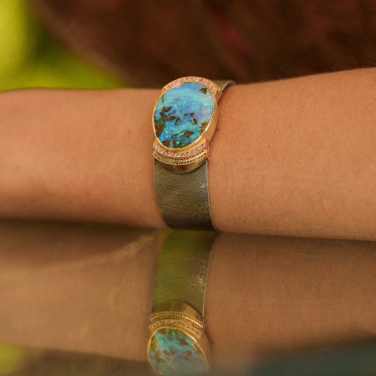 A close-up of a gold-toned cuff bracelet worn on a wrist, featuring a large oval turquoise-blue opal center stone surrounded by small clear stones and a textured hand engraved silver band.