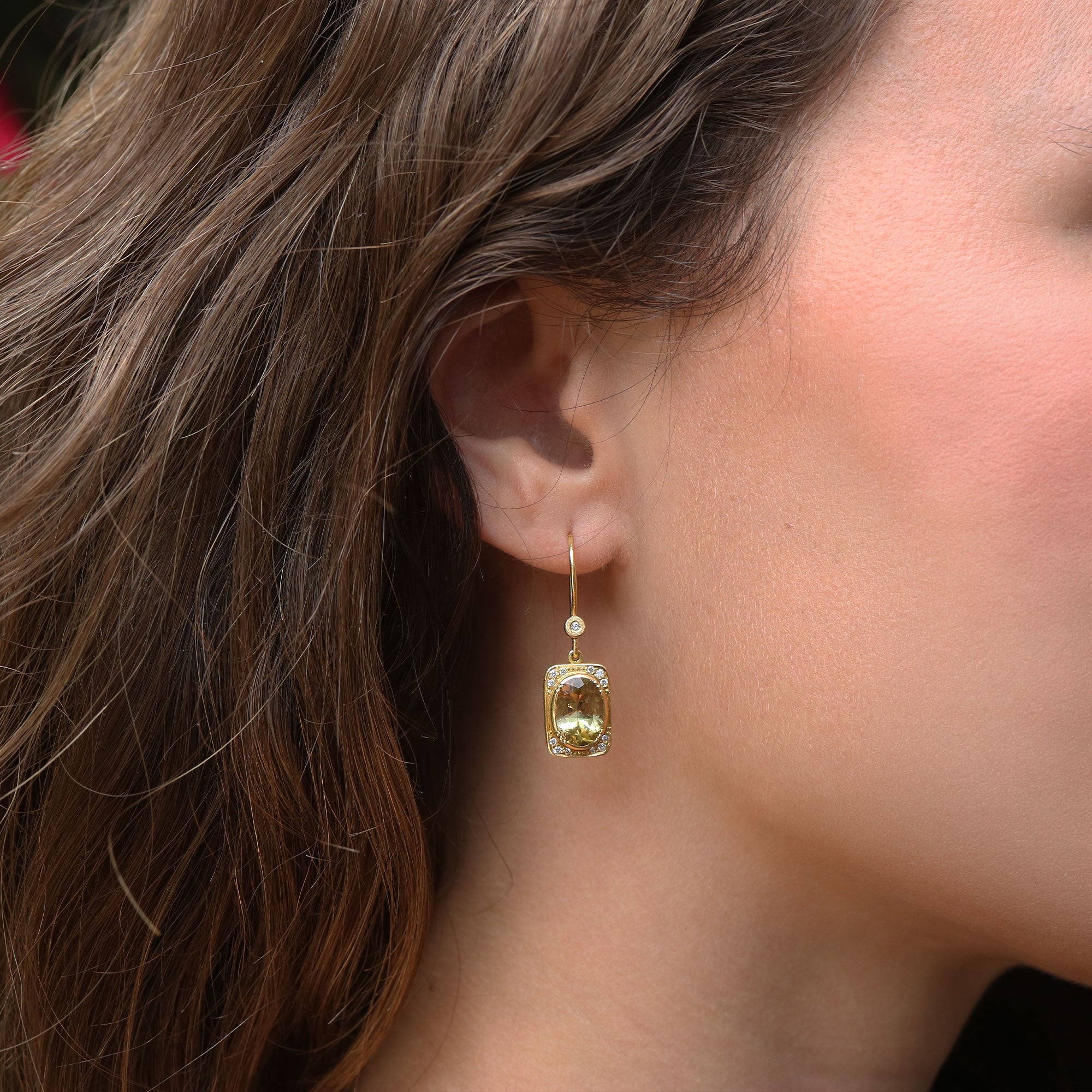 A close-up side profile of a woman wearing a gold dangle earring with a rectangular yellow-green tourmaline gemstone and a small clear accent stone.