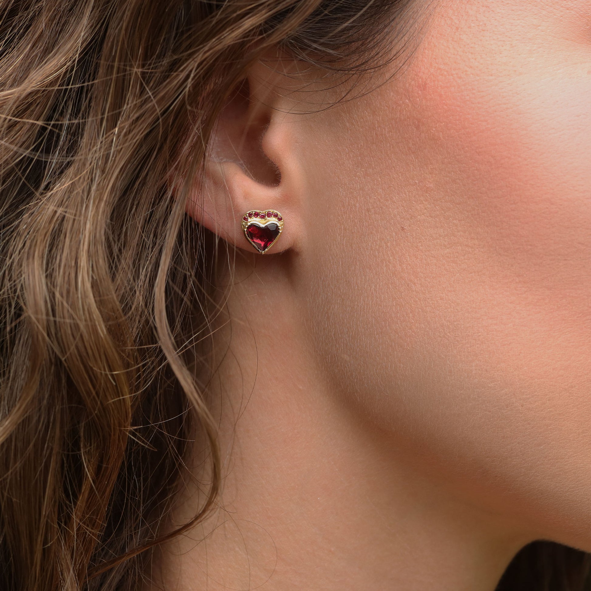 A close-up of a woman's ear wearing a small heart-shaped stud earring with a red ruby heart in a gold-tone setting.