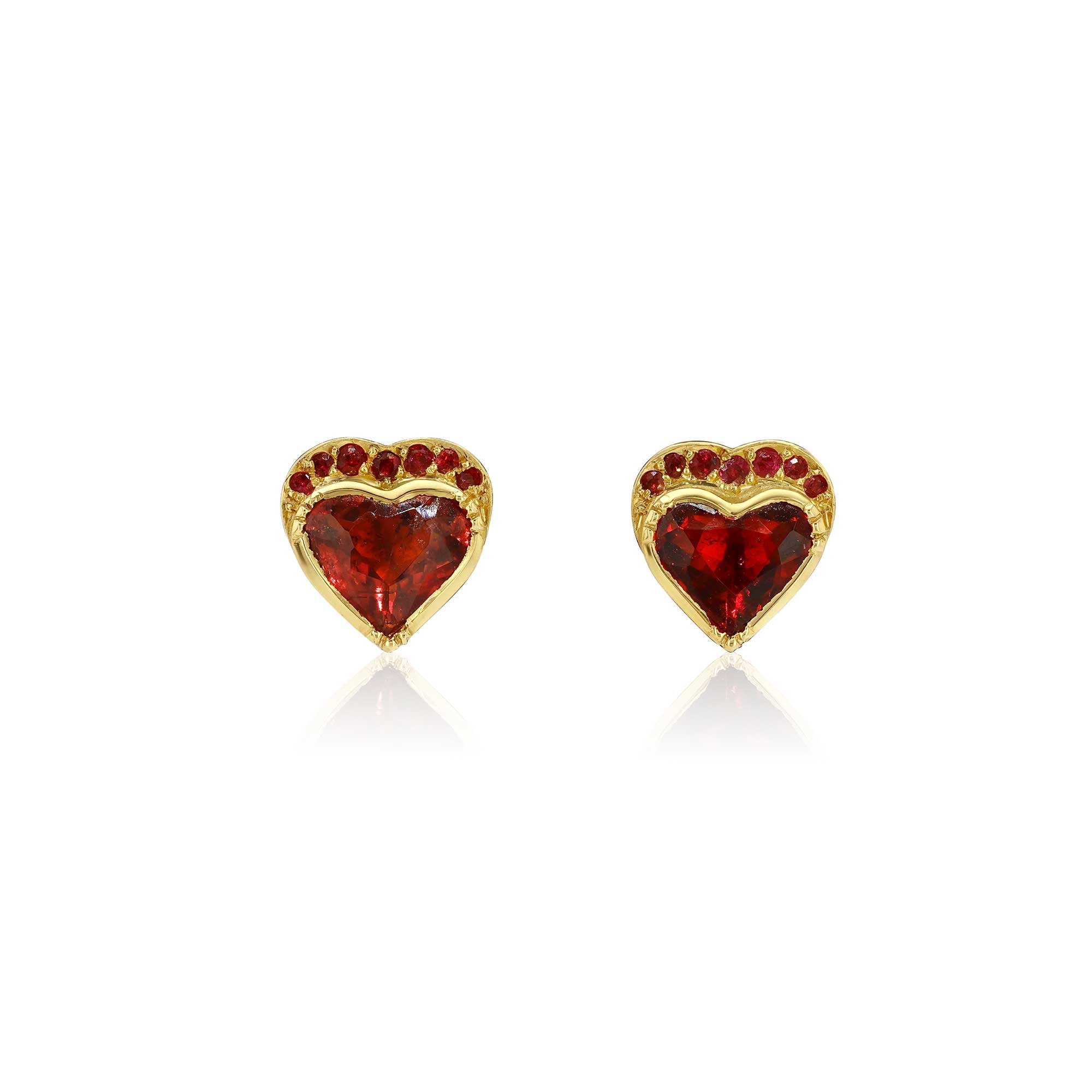 A pair of gold heart-shaped stud earrings featuring a central red heart  ruby gemstone and a border of small red tourmaline stones, shown on a clean white background with a soft reflection.