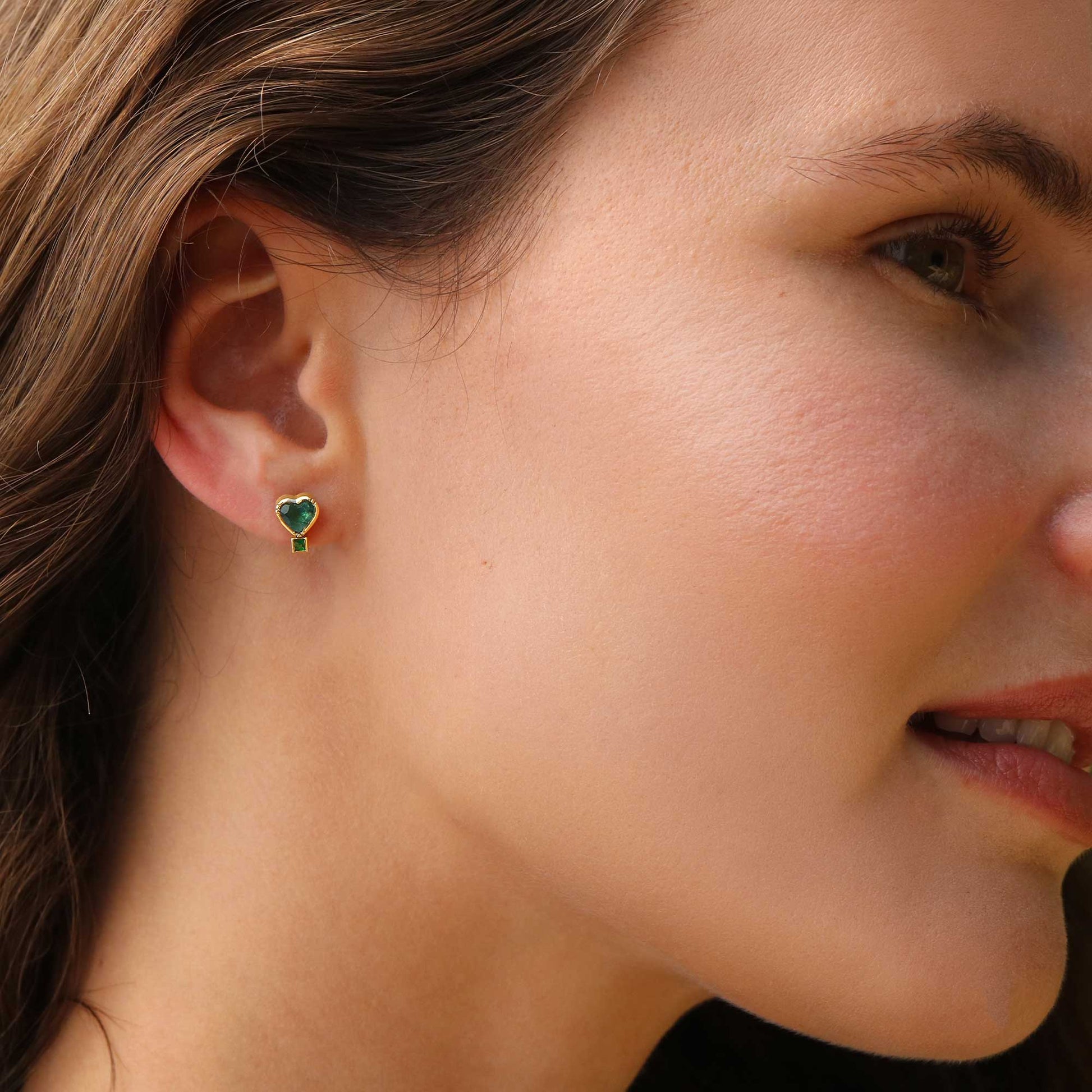 A close-up of a woman's ear wearing a small heart-shaped stud emerald and tourmaline earring.