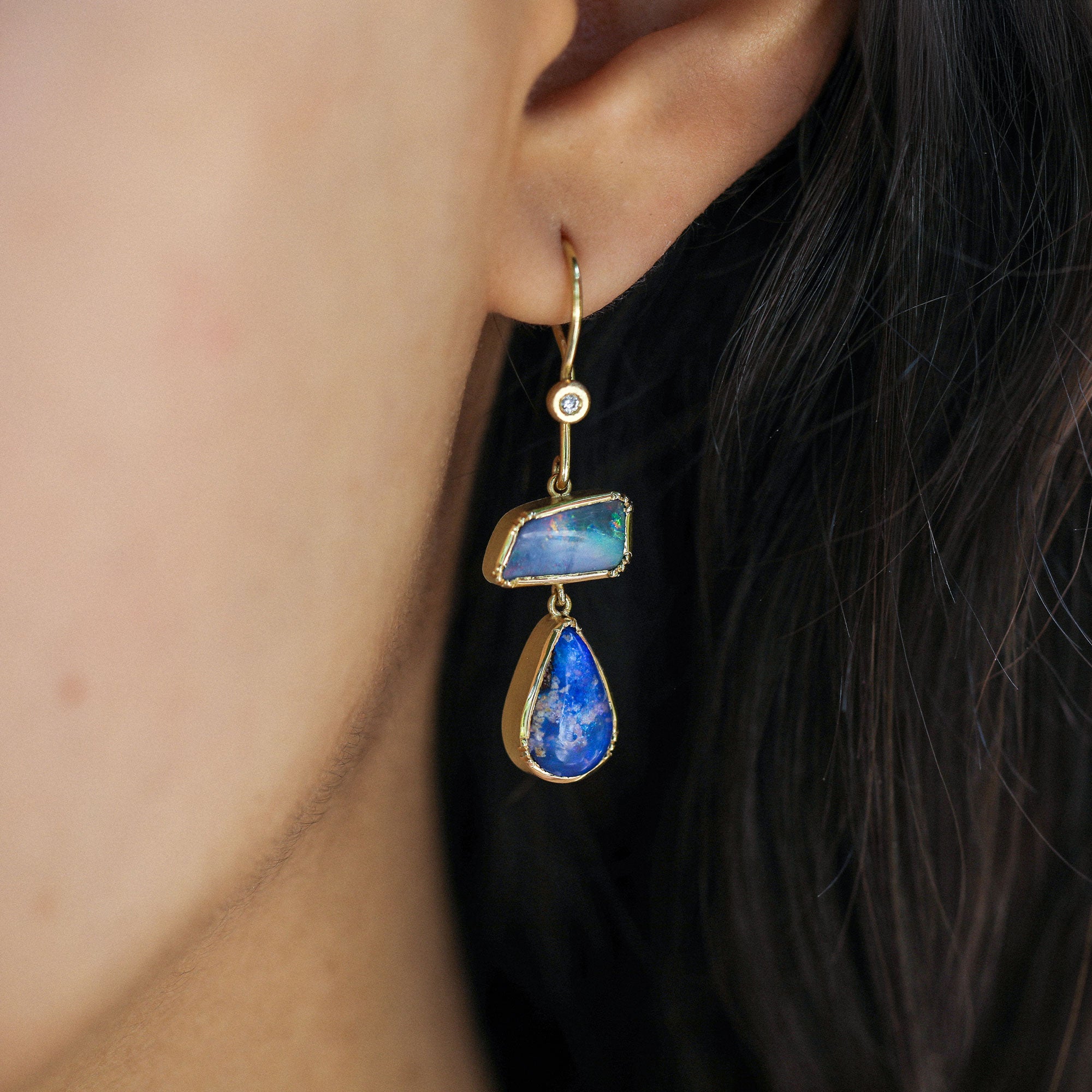 A close-up of a person's ear wearing a gold drop earring with a rectangular opal blue stone and a teardrop-shaped opal blue stone.