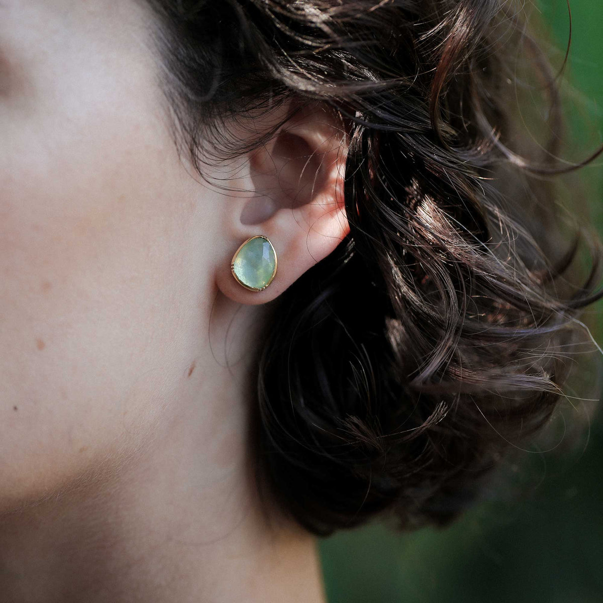 A close-up of a person's ear wearing a gold stud earring with a teardrop-shaped pale blue aquamarine stone.