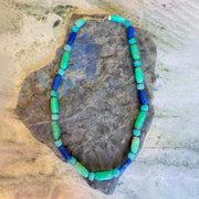 A beaded necklace made of alternating turquoise, light green, and royal blue cylindrical beads with a gold clasp, displayed on a textured gray stone against a light beige stone background.
