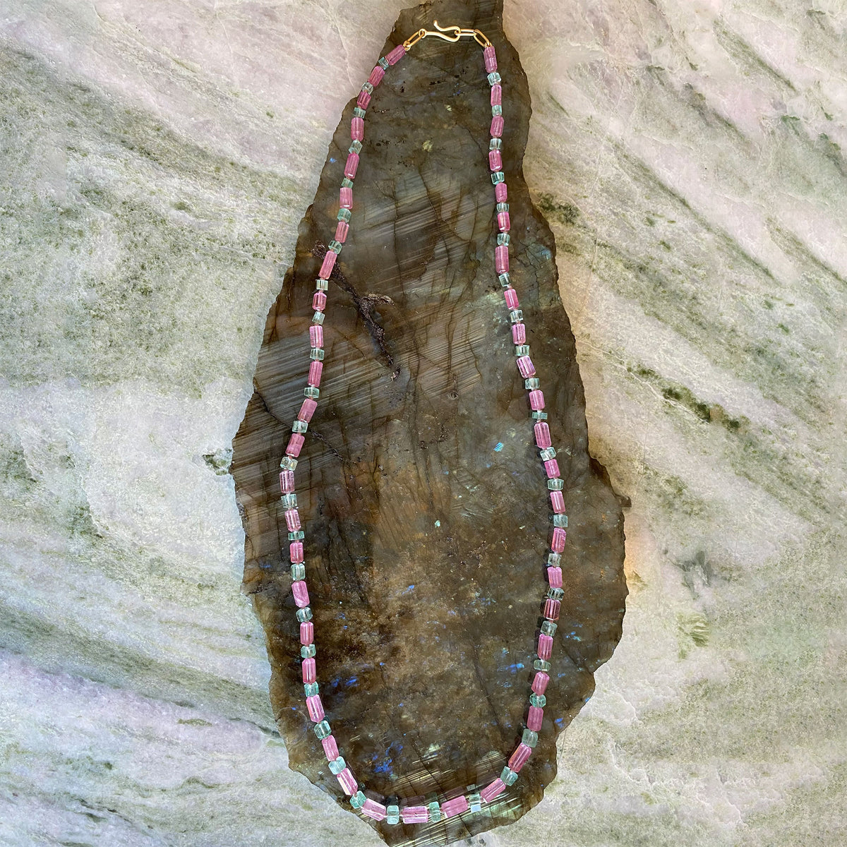 A beaded necklace made of pink cylindrical beads separated by small green and blue beads, displayed on a dark polished stone slab against a light gray textured stone background.