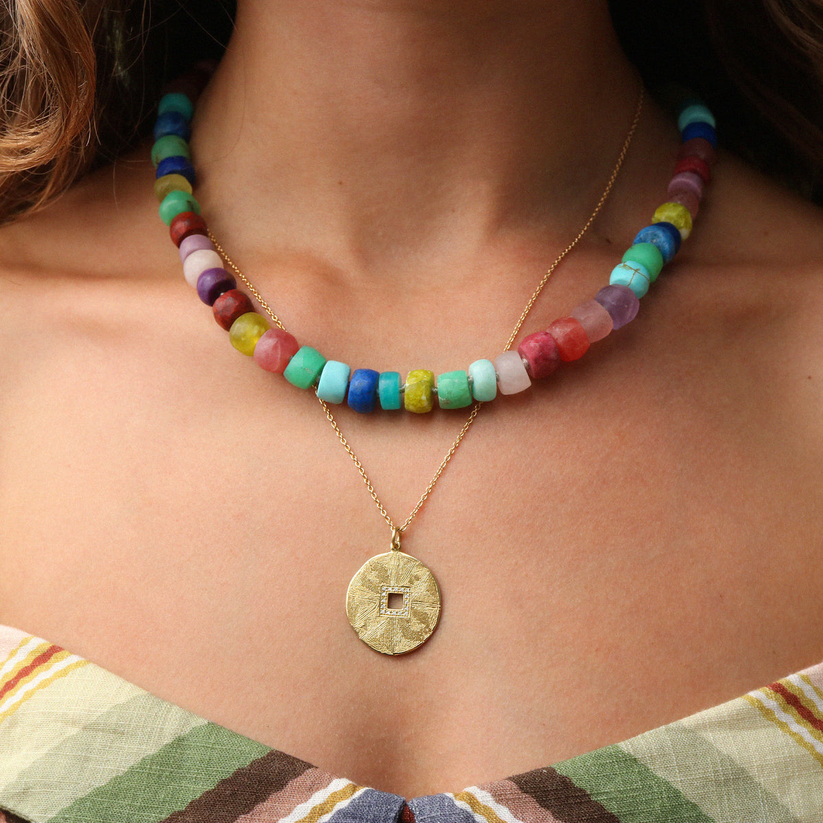 A close-up of a model wearing a multi-colored beaded necklace layered over a gold chain with a round textured coin pendant.