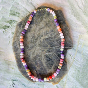 A beaded necklace made of small, irregularly shaped multicolor gemstone beads in shades of purple, pink, orange, red, yellow, and white, with a gold clasp, displayed on a dark polished stone slab over a light textured background.