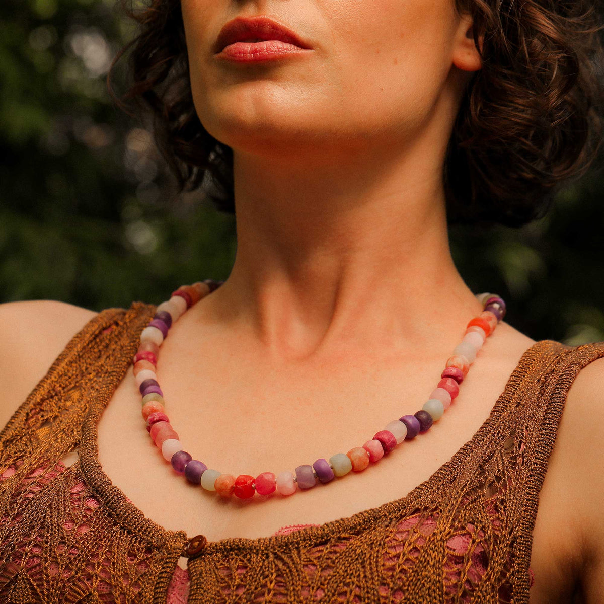 A close-up of a woman wearing a colorful beaded necklace with chunky round beads in shades of pink, purple, orange, and cream, paired with a brown crochet top against a blurred green outdoor background. Bead Necklace designed and made by Brooke Gregson in Los Angeles.