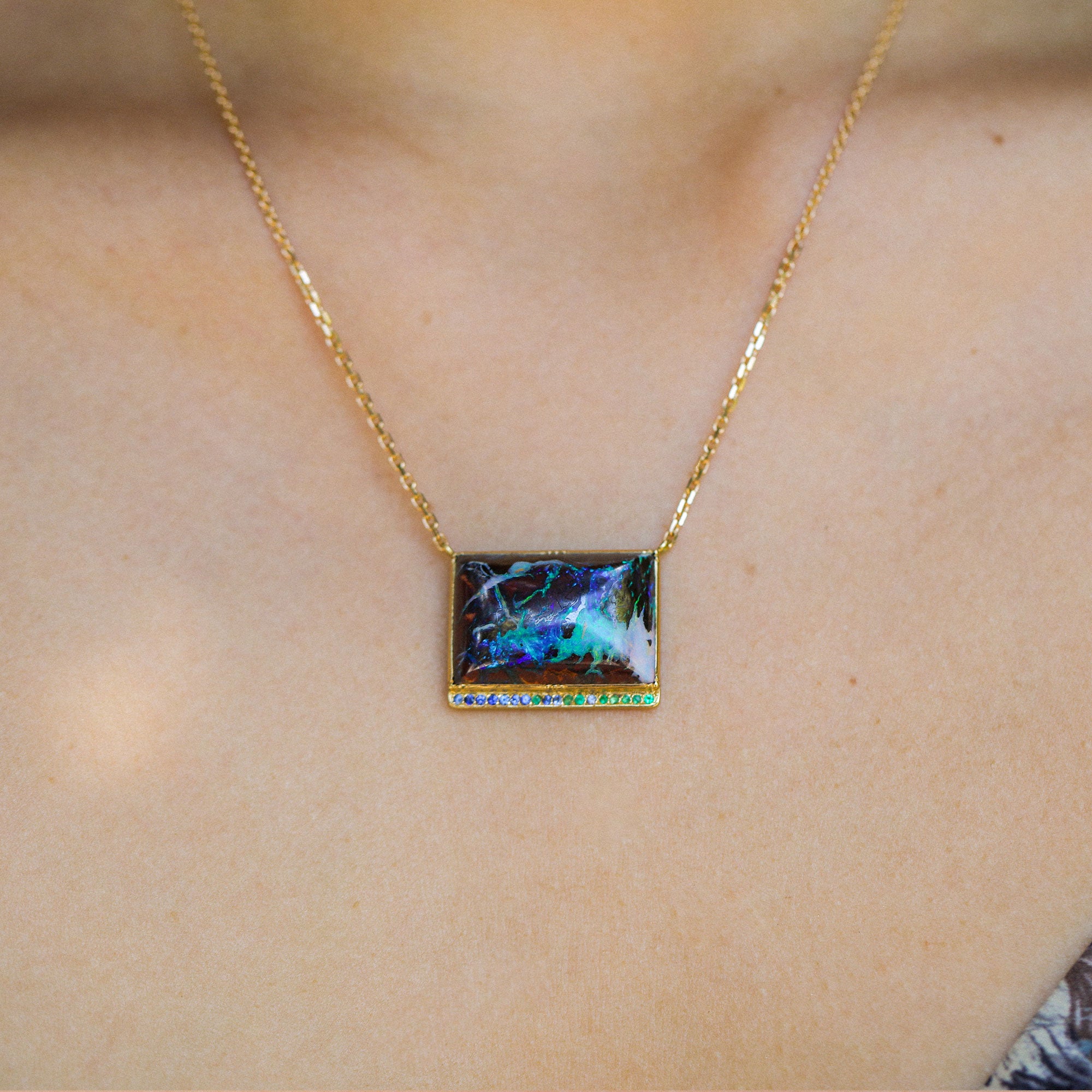 A gold chain necklace with a rectangular pendant featuring a colorful, iridescent opal-like inlay, worn on a person's neck.