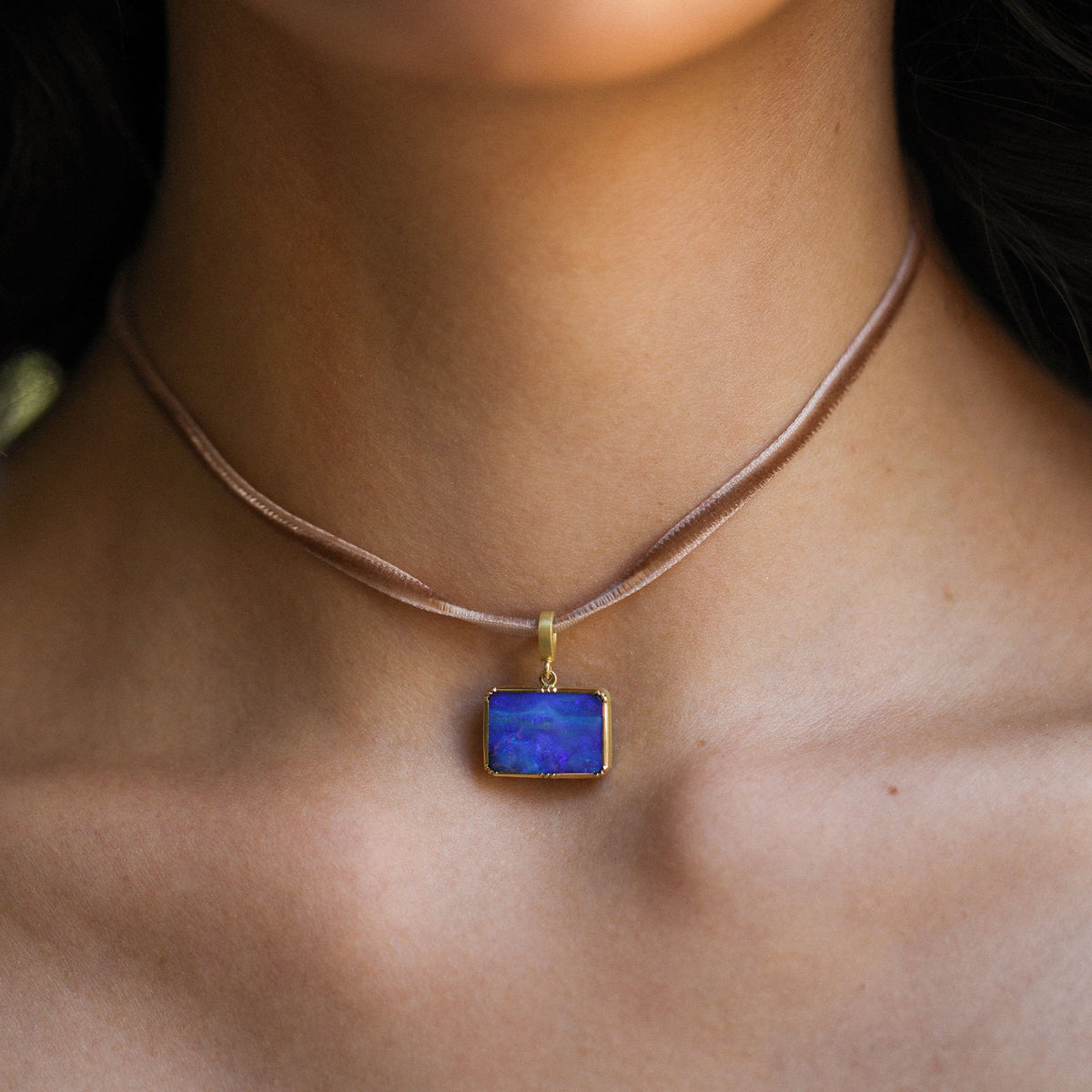 A close-up of a person's neck and chest wearing a delicate brown velvet necklace with a small gold-toned rectangular pendant featuring a deep blue opal stone.