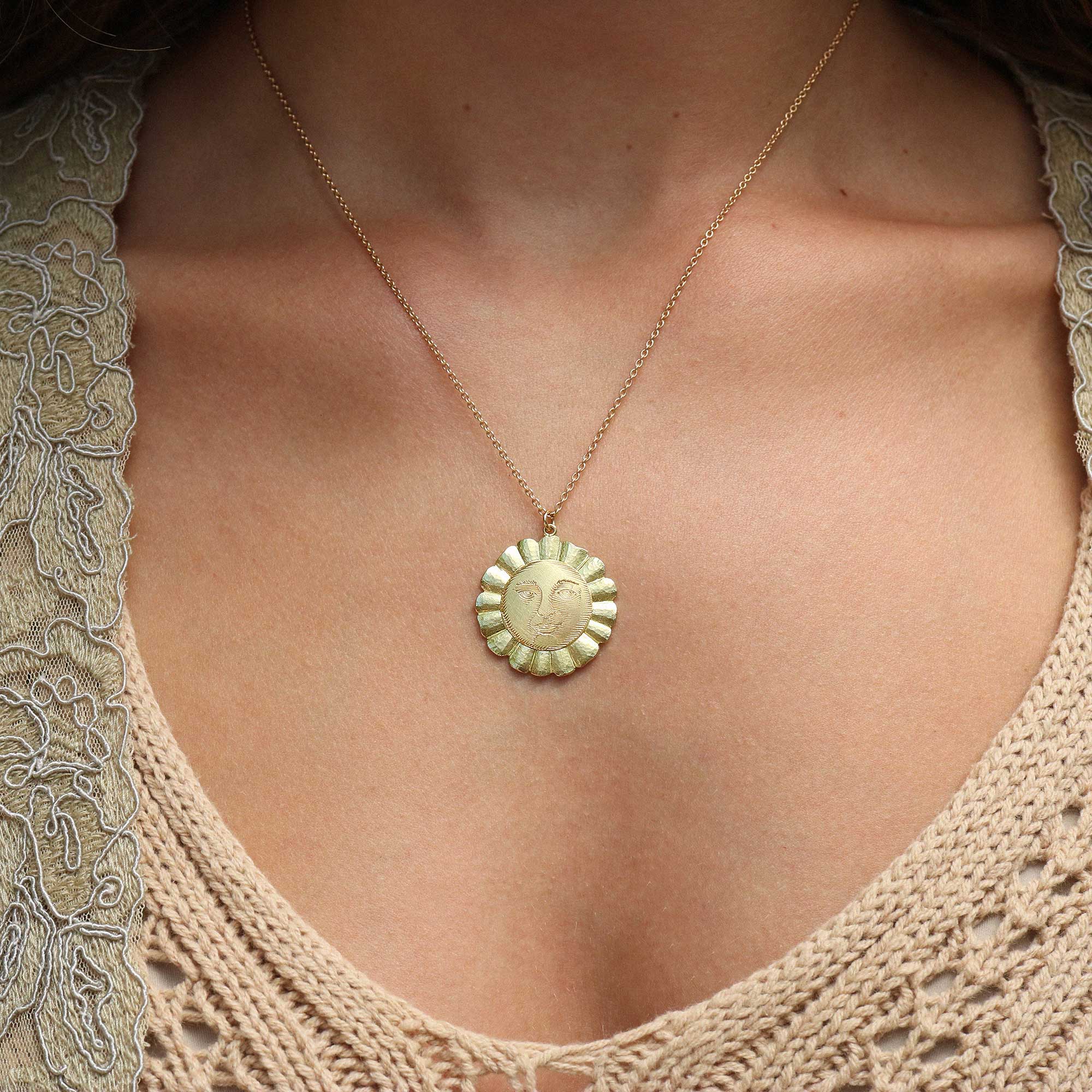 A close-up of a person wearing a delicate gold chain necklace with a round, scalloped gold pendant featuring an embossed sun design.