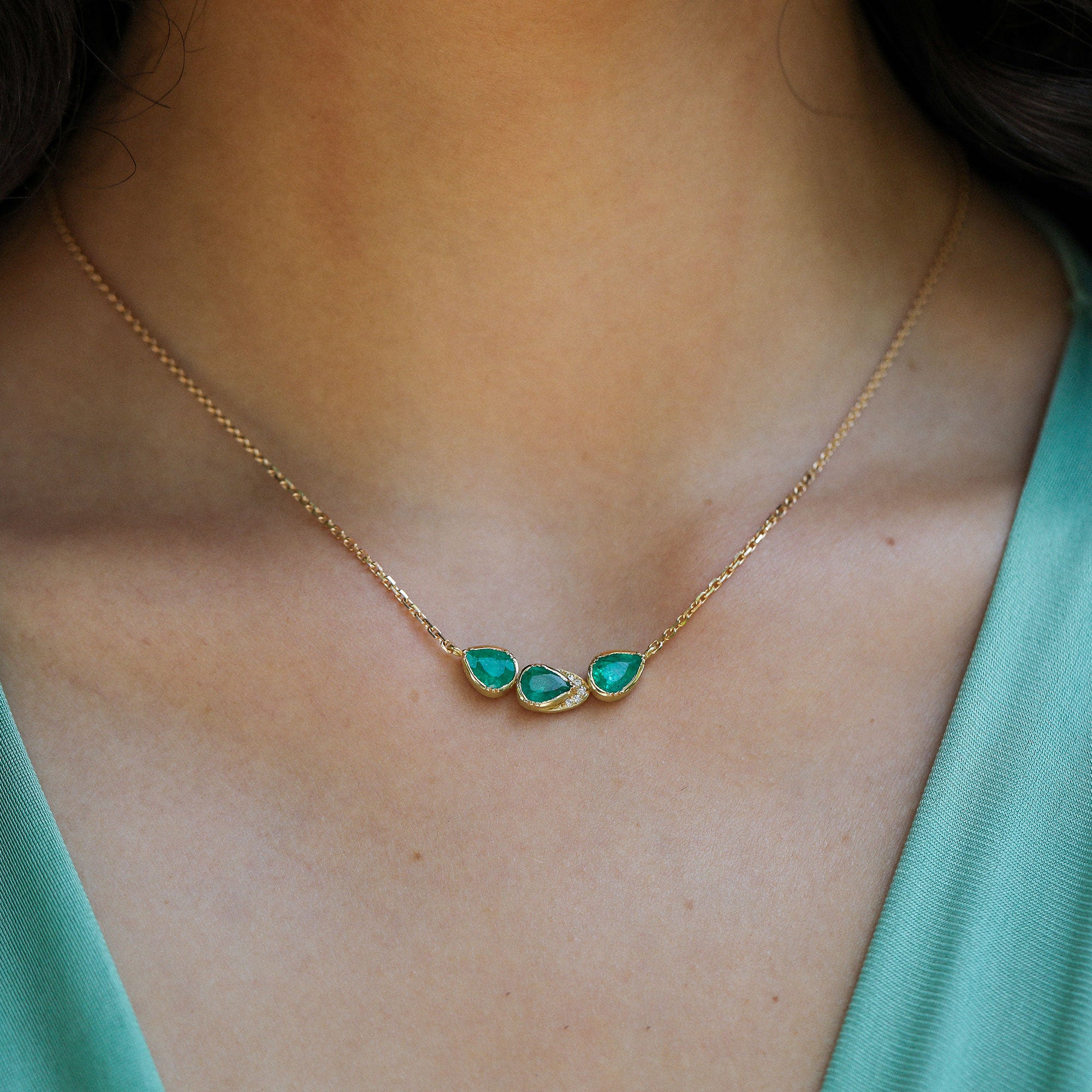A close-up of a person's neck and upper chest wearing a delicate gold chain necklace with three small teardrop-shaped emerald stones set in gold.