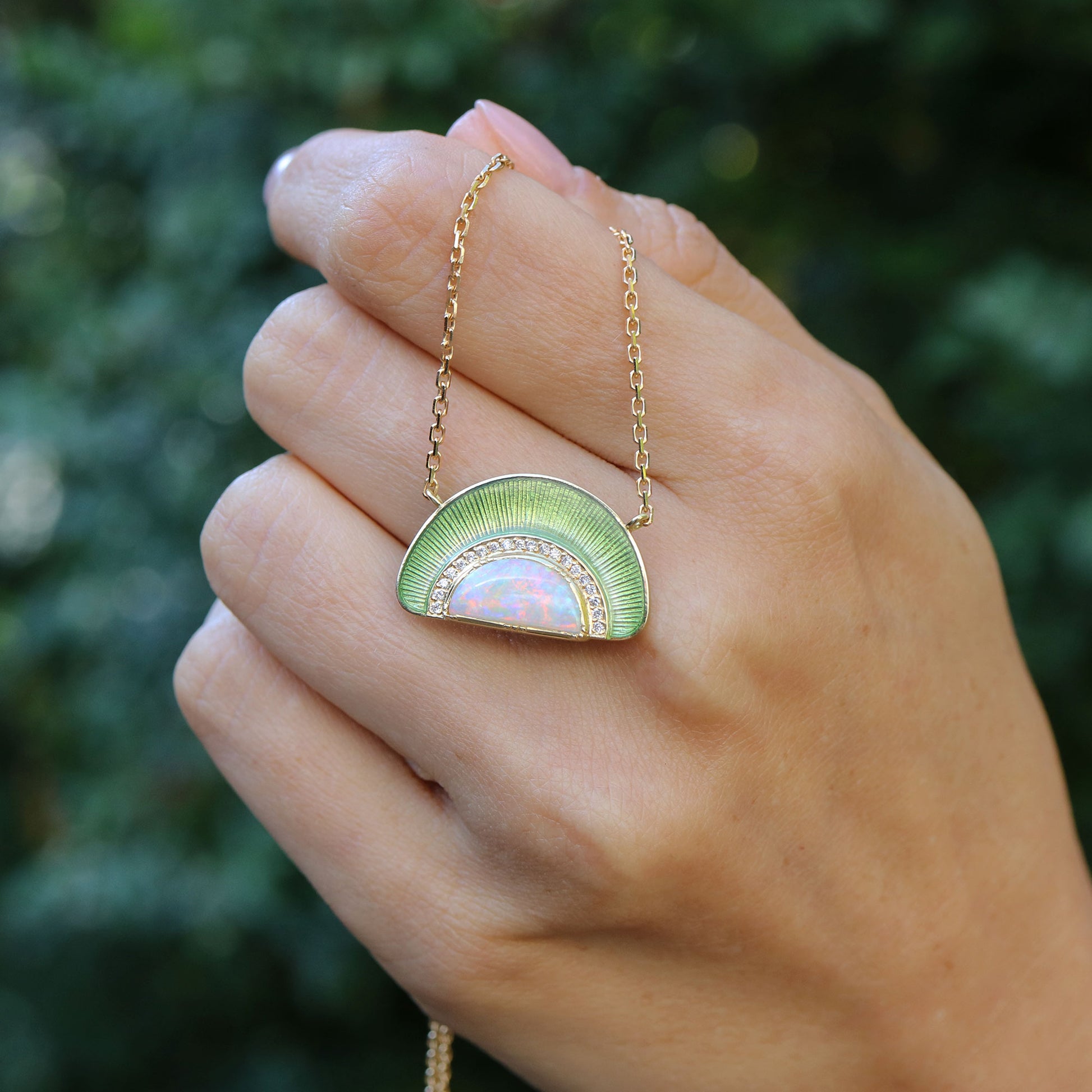 Hand holding a 18k gold necklace with a pendant with opal, white diamonds and sage green enameling by Brooke Gregson.
