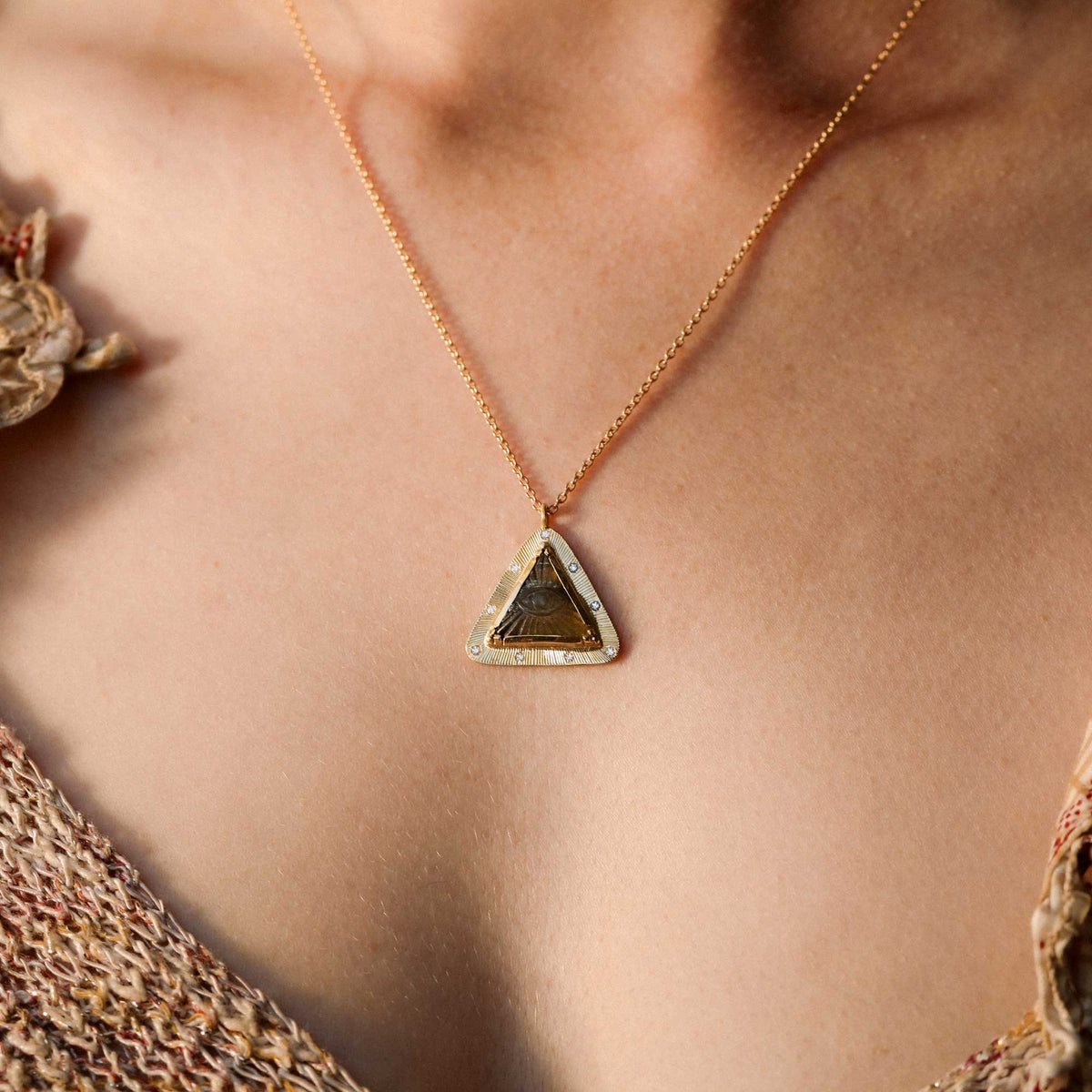 A close-up of a person wearing a delicate yellow gold chain necklace with a triangular eye pendant. The pendant features a textured outer edge surrounded by a border of small diamonds, set against warm skin tones.