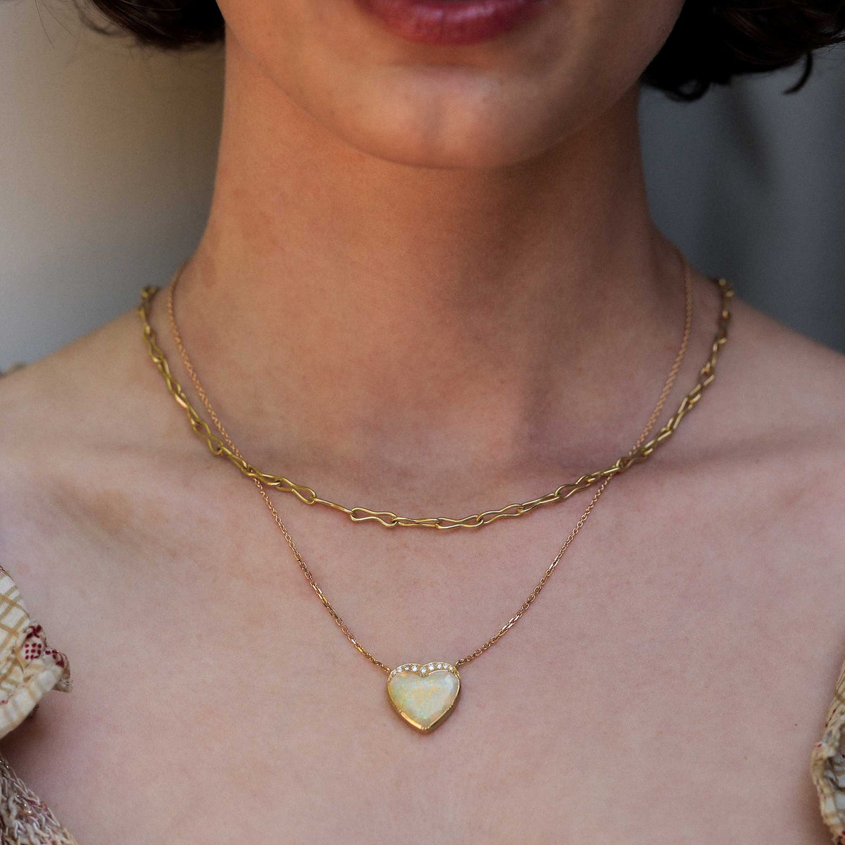 A close-up of a model's neck and chest wearing two layered gold necklaces: a thicker twisted chain and a delicate chain with a heart-shaped opal pendant.