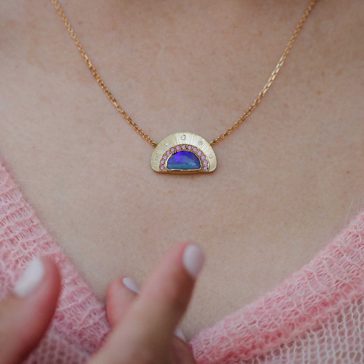 A close-up of a gold necklace with a semi-circular pendant featuring a blue iridescent opal center and small pink garnet stones, worn on a person's neck above a pink knit top.