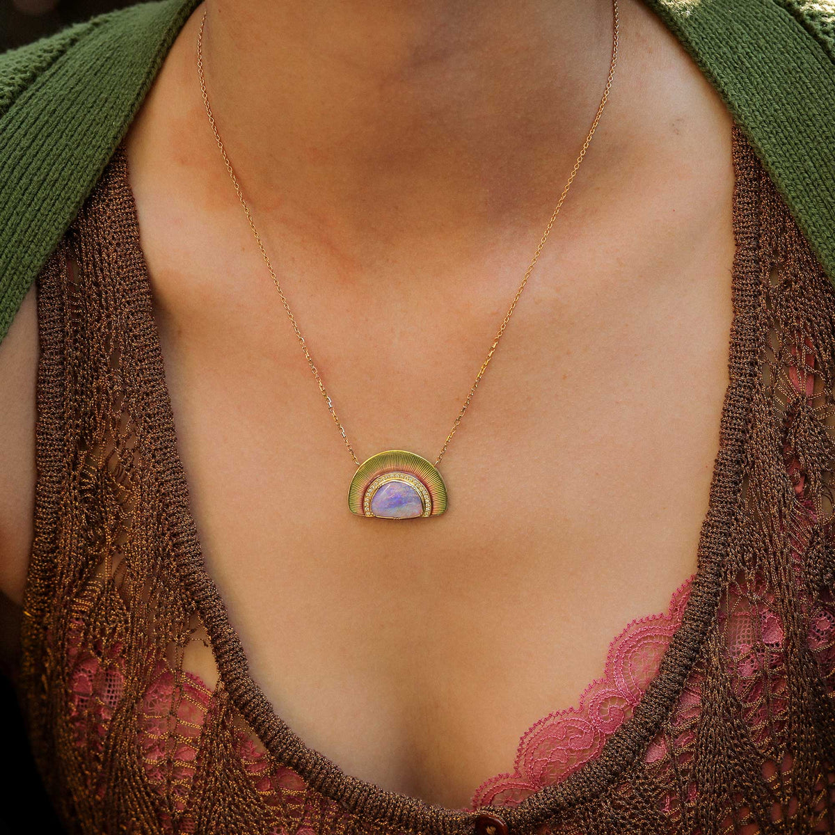 A close-up of a person wearing a delicate gold chain necklace with a small rainbow enamel pendant. The pendant features concentric arches in olive green, gold, and pink, with a smooth lavender-purple opal center with white diamond pave. designed in london by jewelry designer brooke gregson.