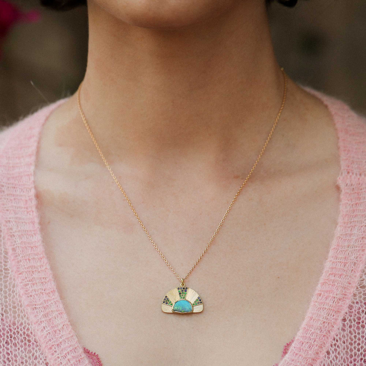 A close-up of a person wearing a delicate 18k gold chain necklace with a small fan-shaped hand engraved pendant featuring a turquoise blue opal center and green tsavorite and emerald details, paired with a pink knit top.