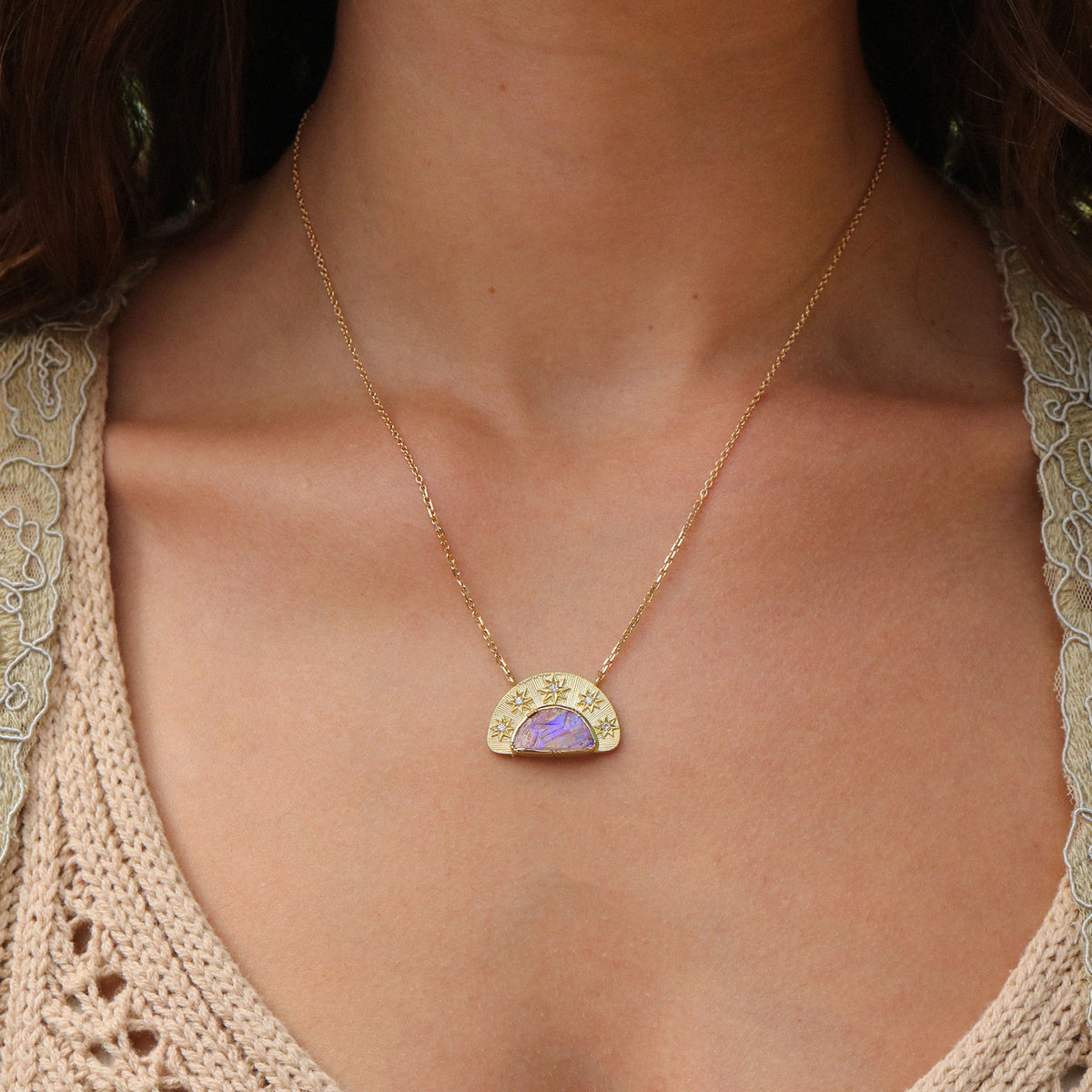 A gold pendant necklace featuring a semi-circular gold frame with small star details and tiny diamond stones, surrounding a central purple opal inlay. The necklace is displayed on a persons neck with a lace white top and dark hair.