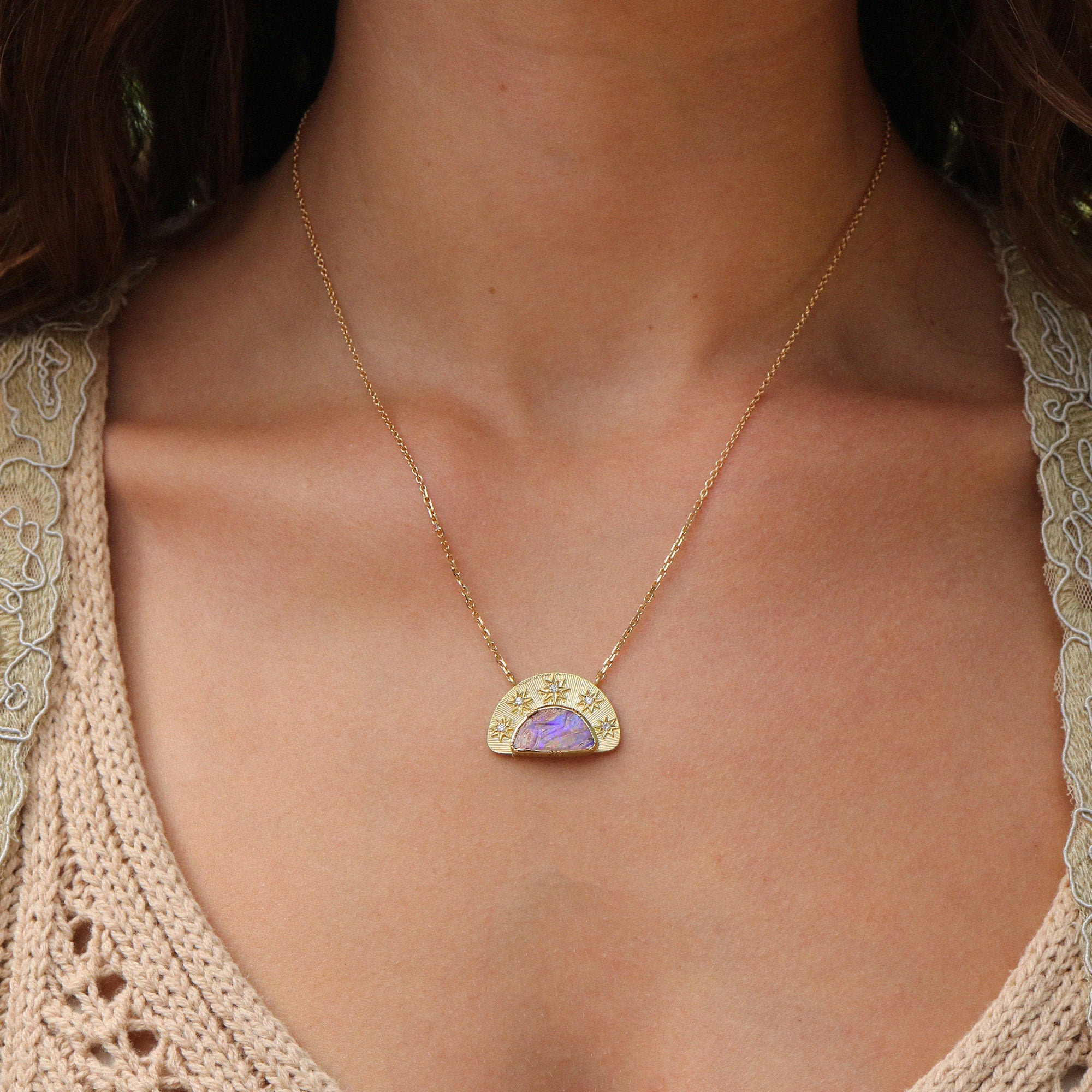 A gold pendant necklace featuring a semi-circular gold frame with small star details and tiny diamond stones, surrounding a central purple opal inlay. The necklace is displayed on a persons neck with a lace white top and dark hair.