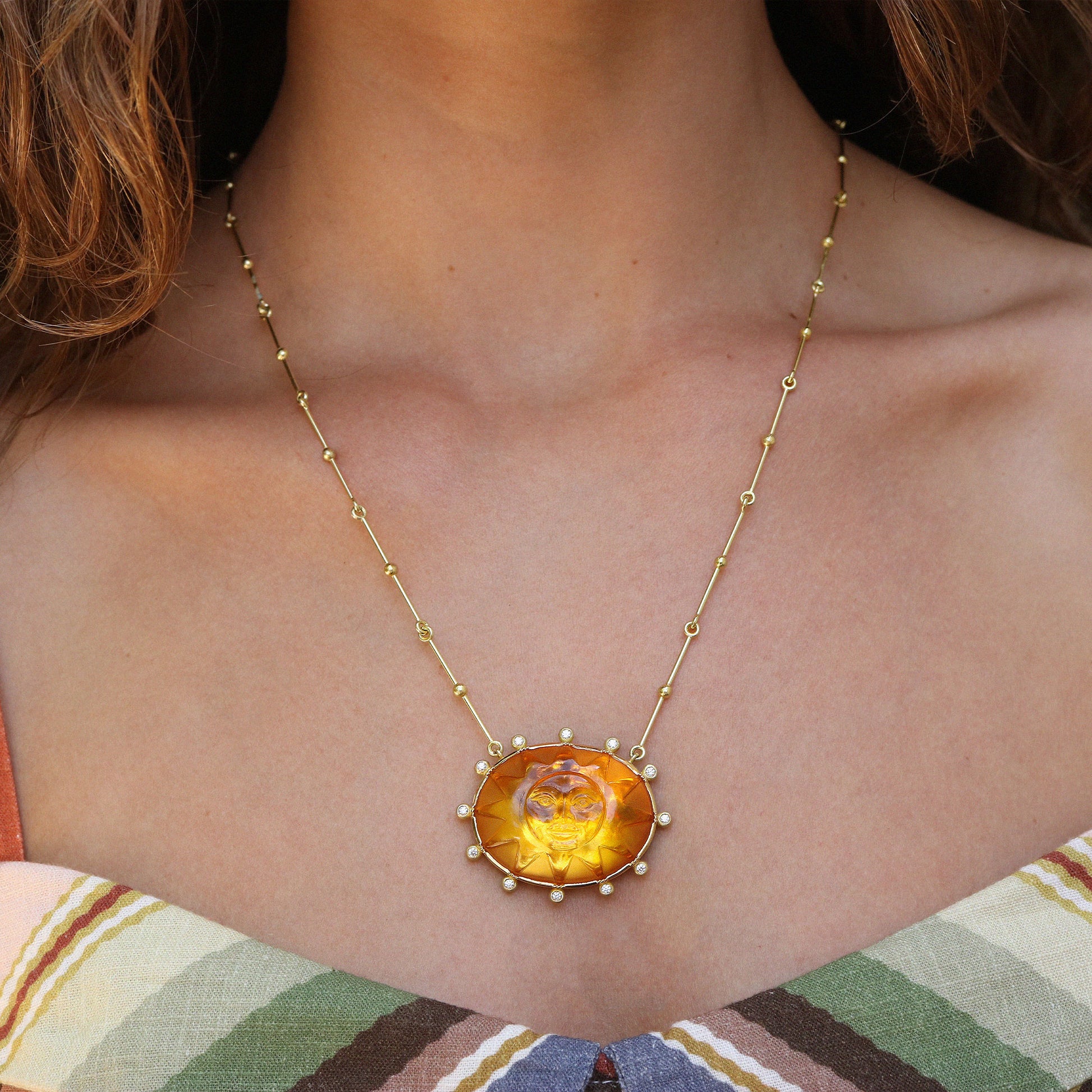 A close-up of a woman wearing a gold necklace with a large oval citrine pendant featuring a carved face.
