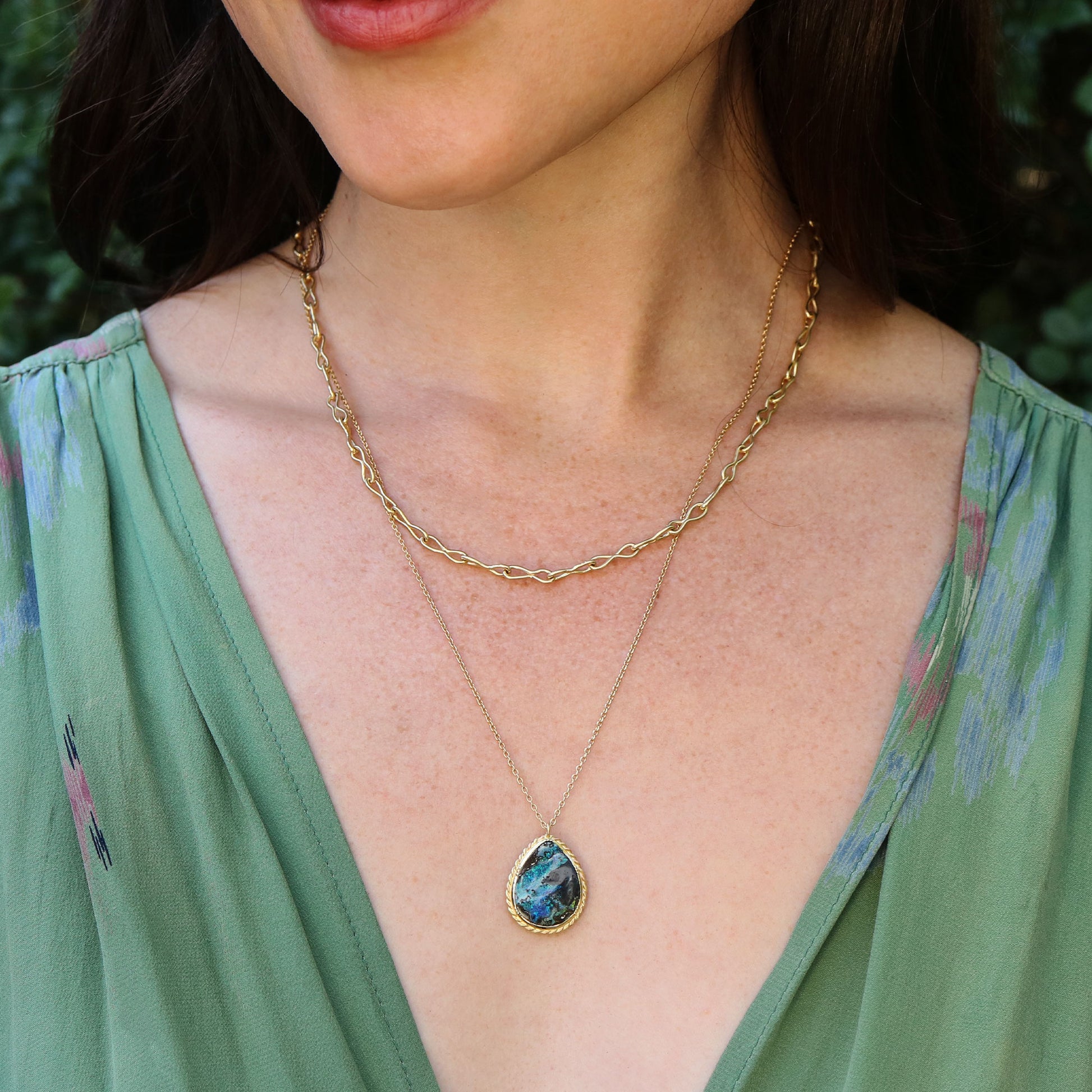 Close-up of a woman wearing a gold necklace with a boulder opal pendant, against a blurred natural background.