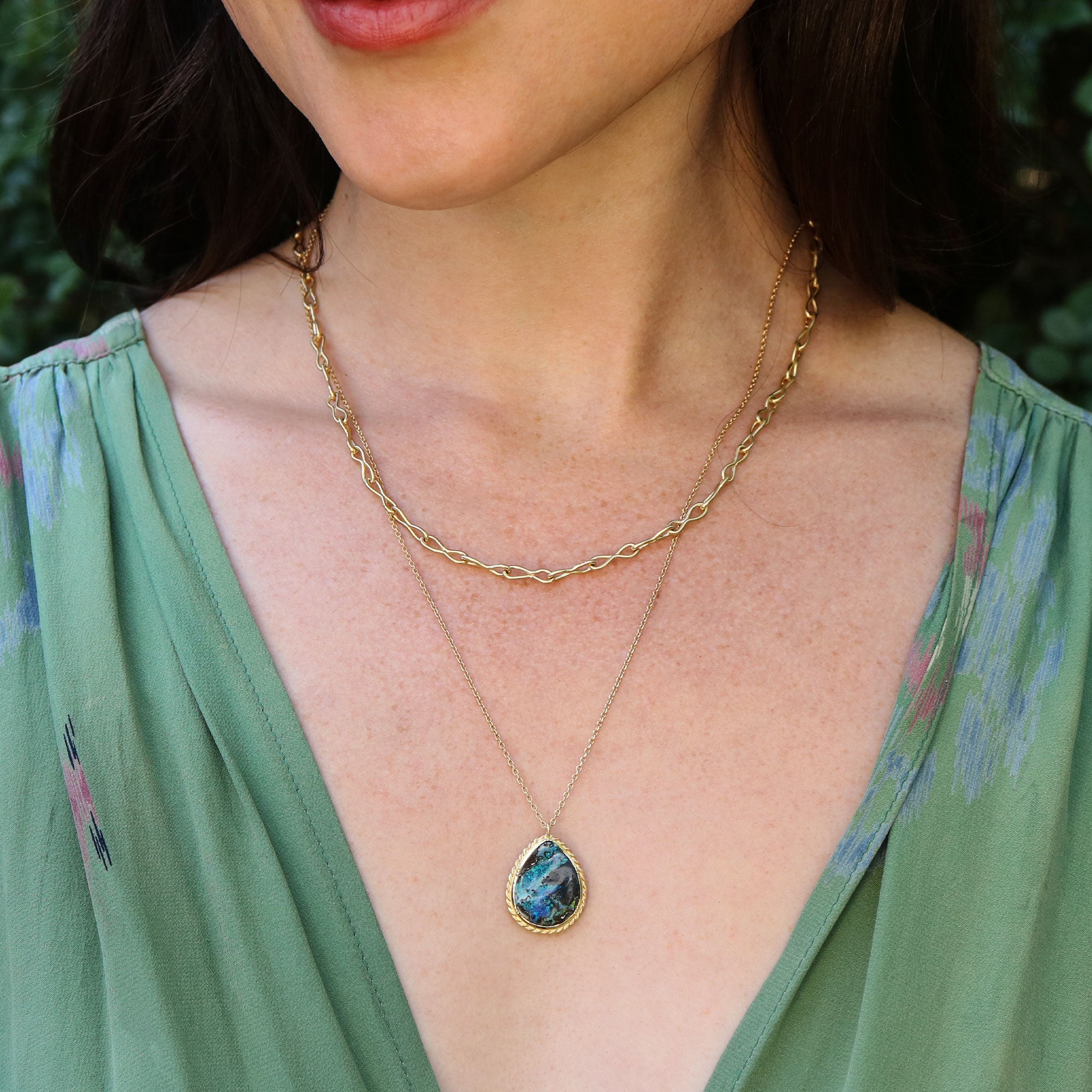 Close-up of a woman wearing a gold necklace with a boulder opal pendant, against a blurred natural background.