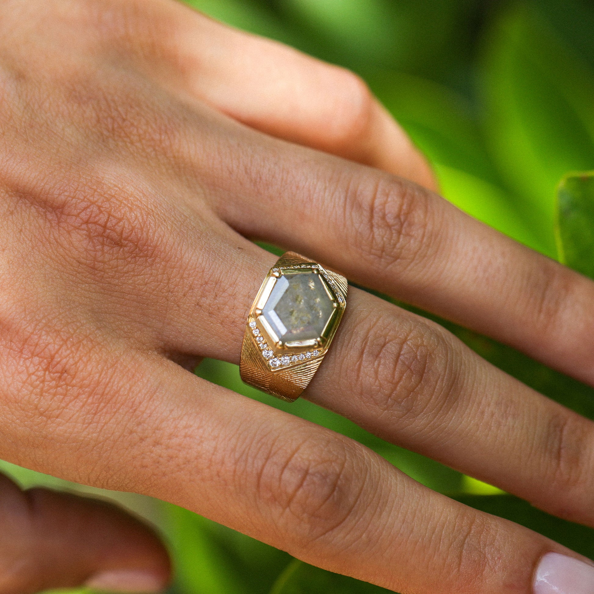 A gold ring with a faceted clear raw diamond gemstone and small pavé diamond accents, shown worn on a person's finger against a blurred green background.
