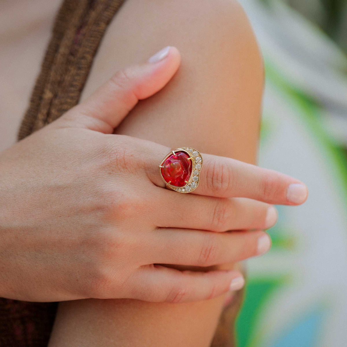 A close-up lifestyle shot of an 18k yellow gold brooke gregson ring with a large red cabochon fire opal gemstone and small diamond accent stones, worn on a person's finger.