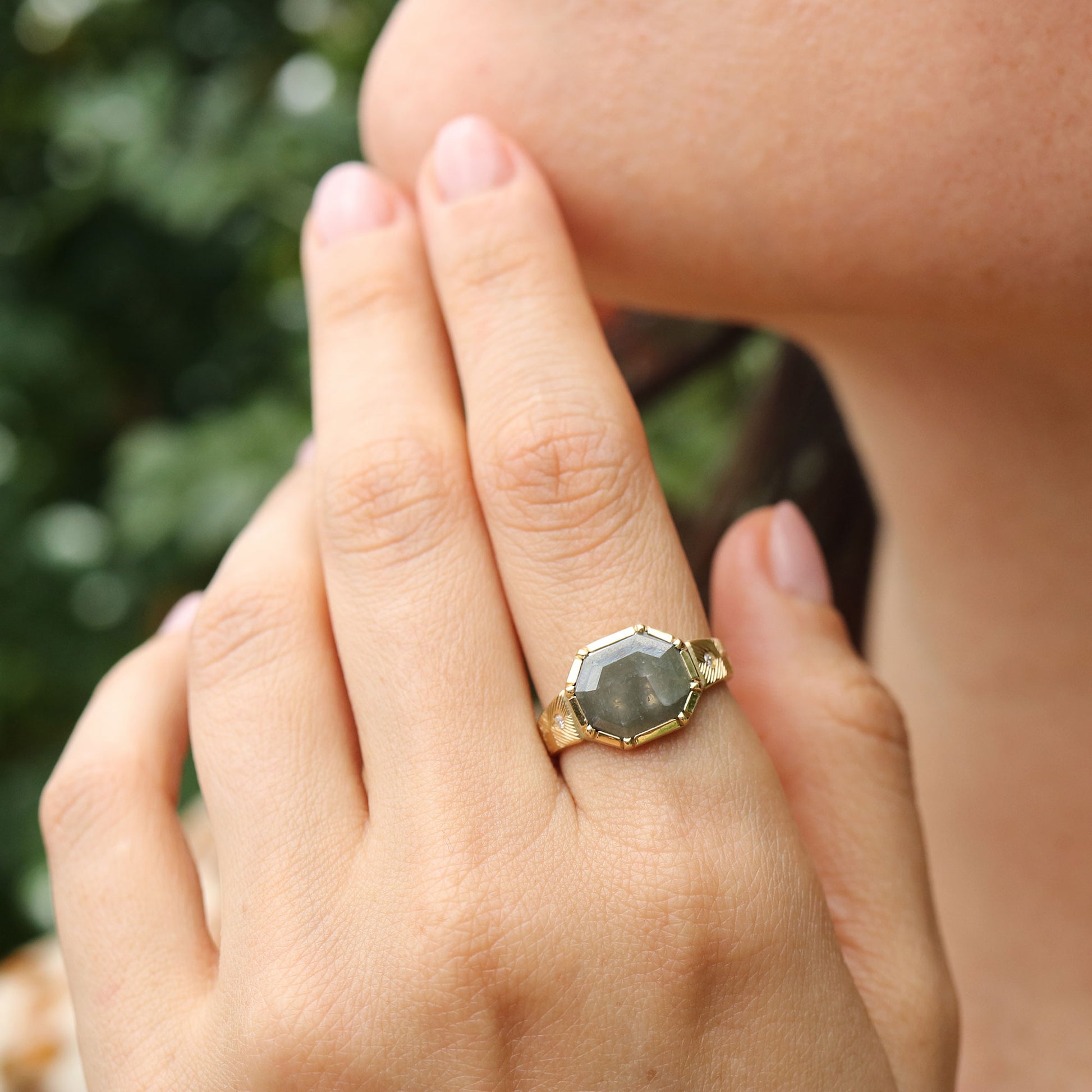 Close-up of a hand wearing a gold ring with a raw diamond gemstone, blurred green background
