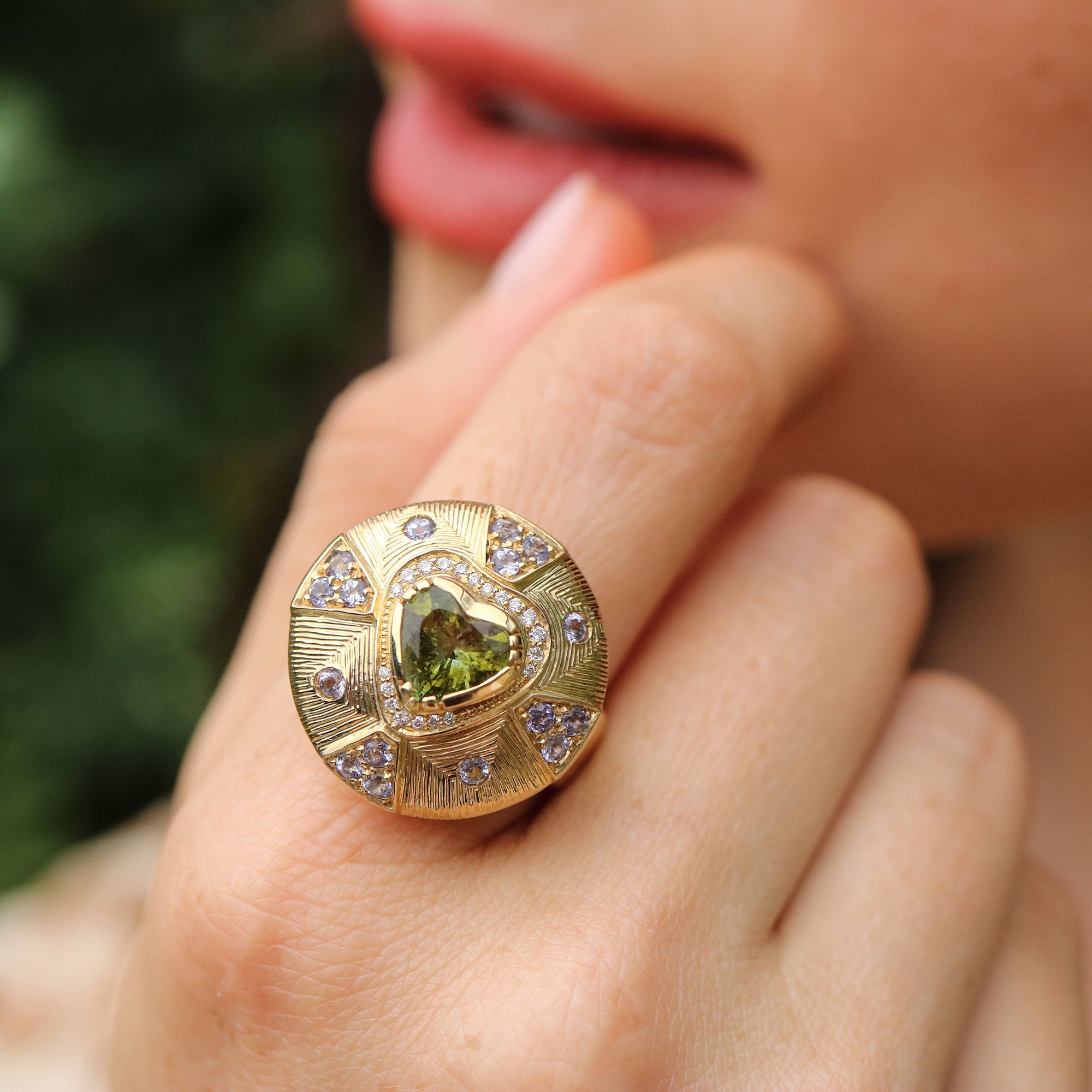 Gold ring with a green tourmaline gemstone and purple tanzanite worn on a finger against a blurred background