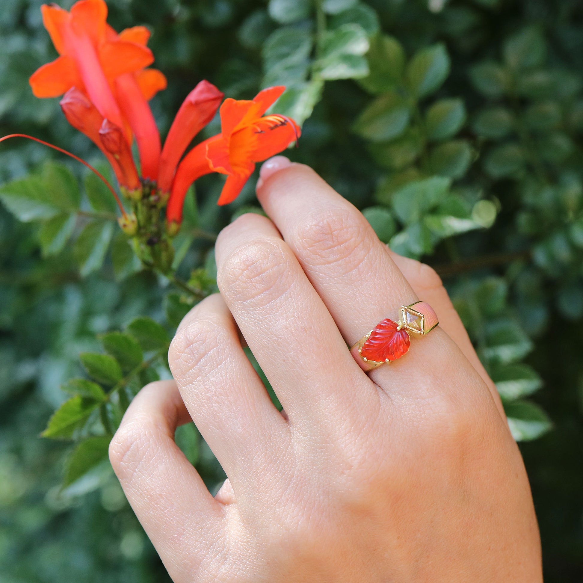 Hand wearing a red fire opal and gold ring with a floral design, holding a red flower against a green leafy background.
