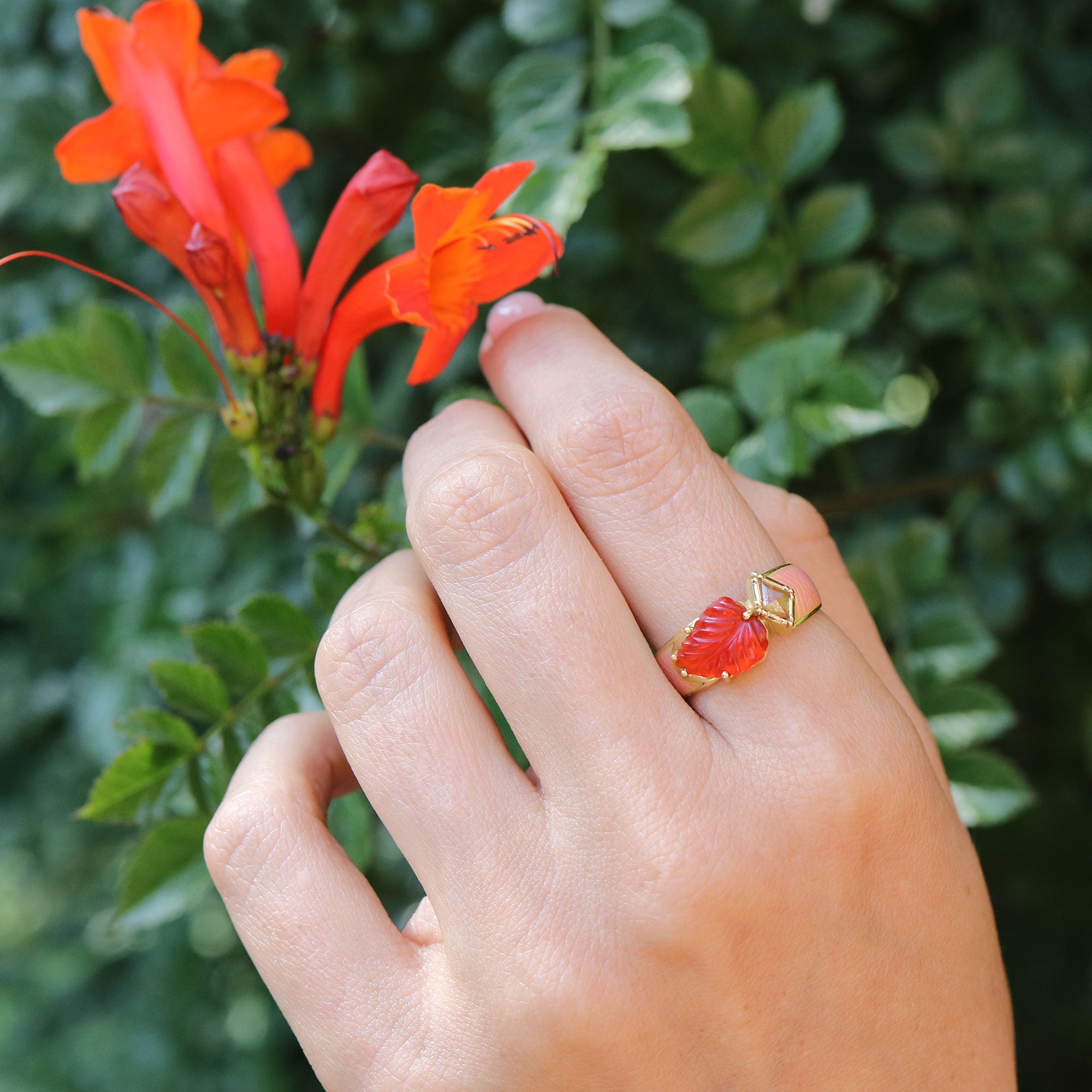 Hand wearing a red fire opal and gold ring with a floral design, holding a red flower against a green leafy background.