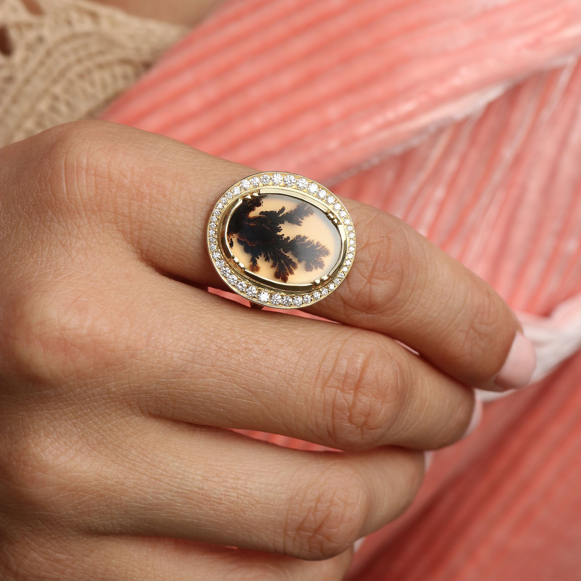 A gold oval ring with a central black and beige tree-of-life dendritic agate, surrounded by small clear stones, worn on a hand against a pink striped background.
