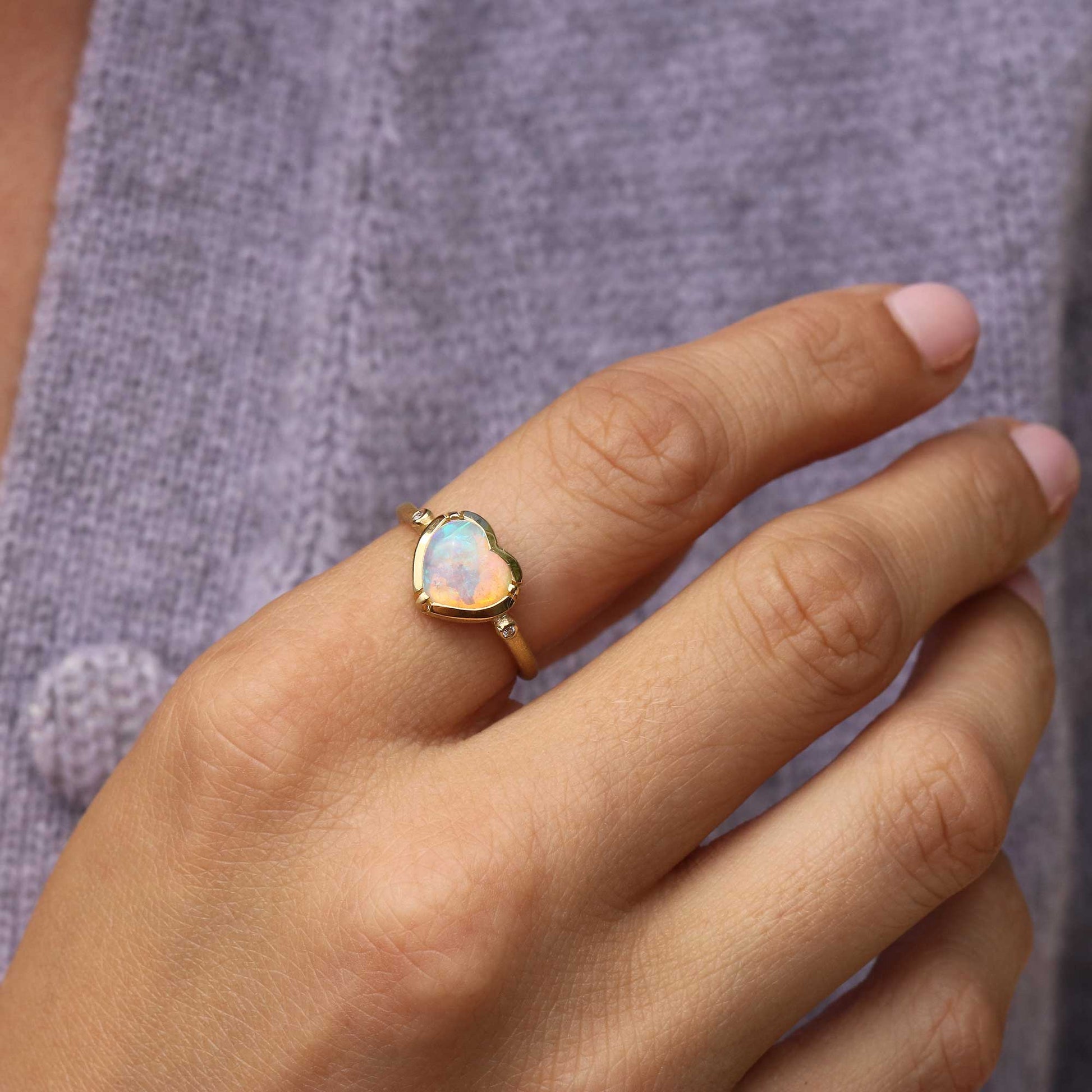 A gold heart-shaped ring with an iridescent opal center, worn on a person's ring finger against a textured purple knit background.
