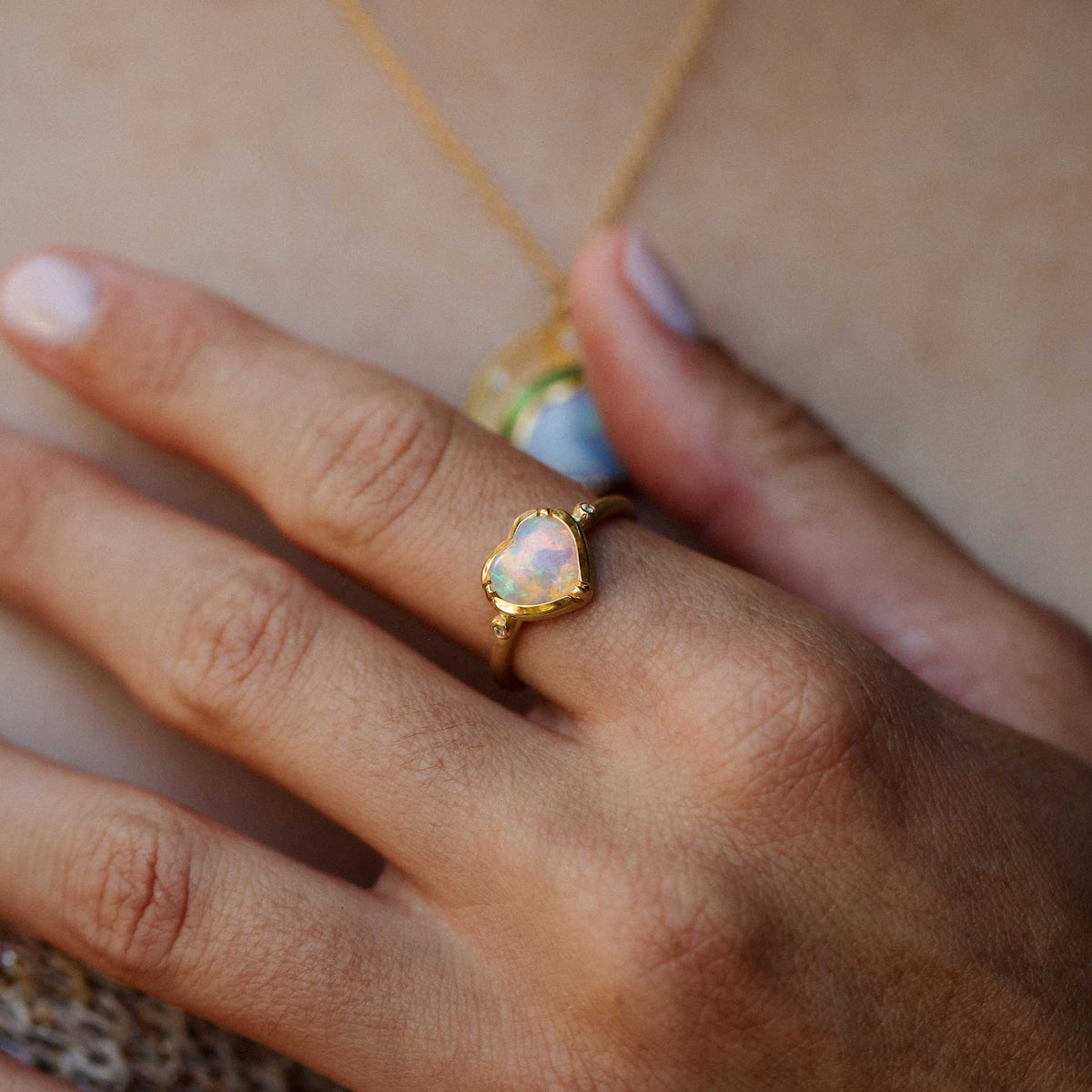 A gold ring with a heart-shaped australian opal gemstone worn on a person's ring finger, shown in a close-up lifestyle shot against a warm beige skin background.