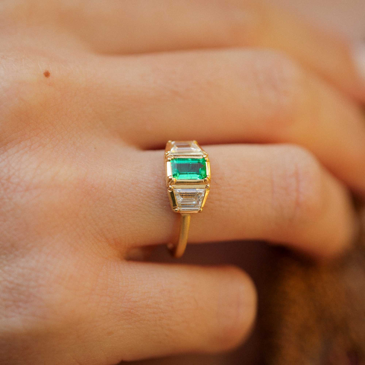 A close-up of an 18k gold ring with a rectangular green emerald-cut center emerald stone and baguette-cut diamond side stones, worn on a person's finger.