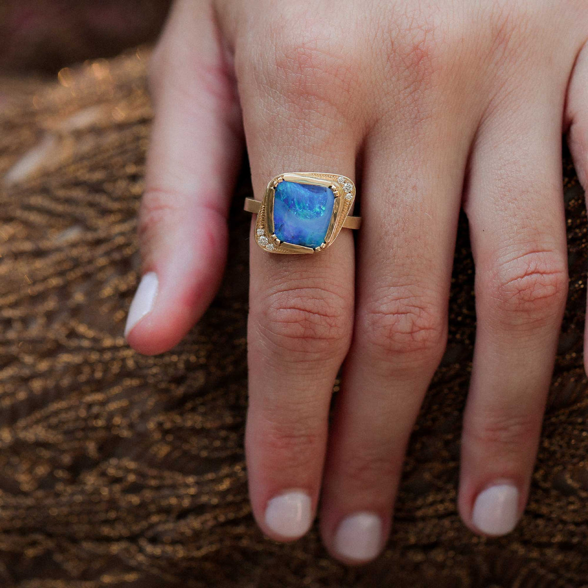 A gold ring with a square blue boulder opal center stone and small diamond accent stones, worn on a person's finger against a textured brown background.