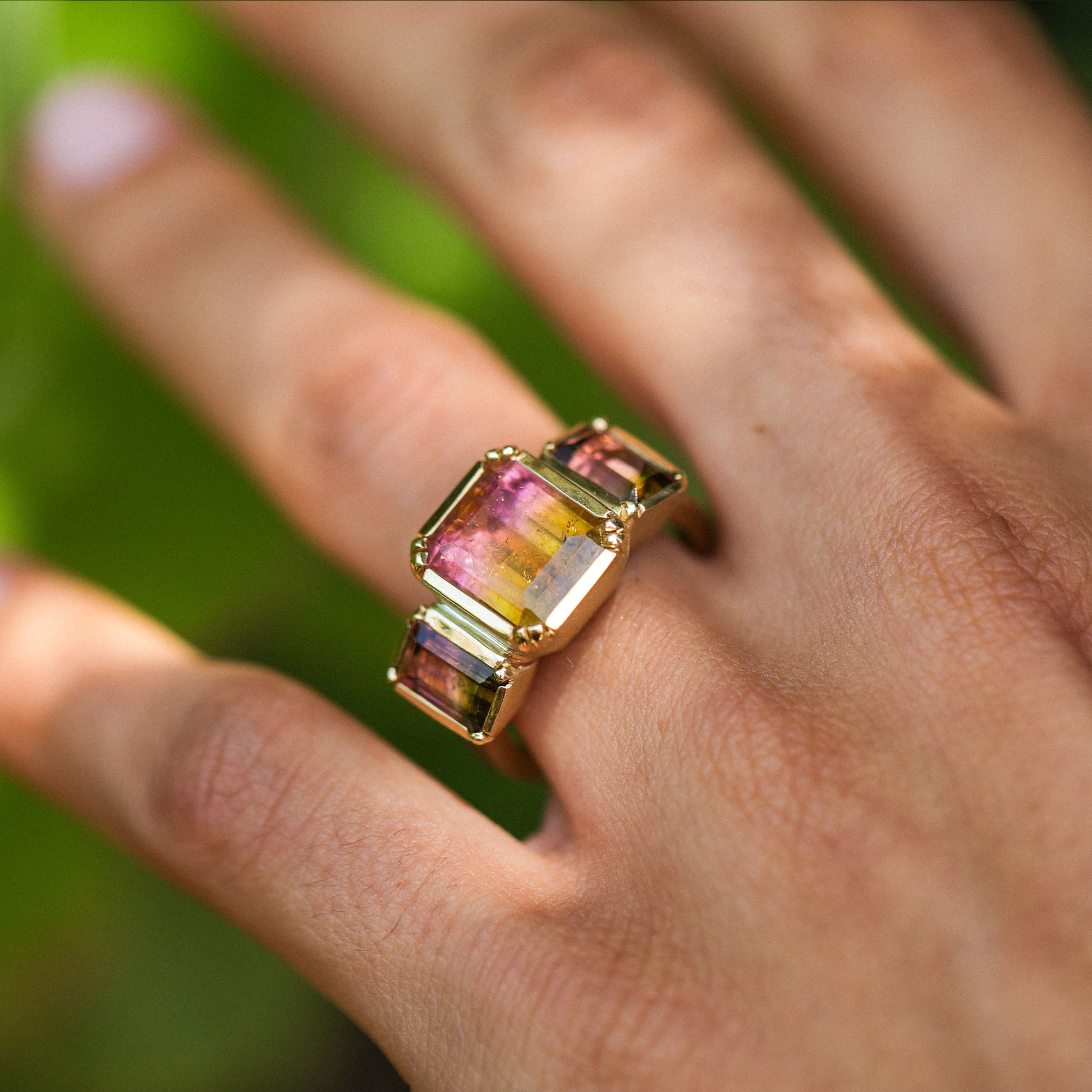 A gold ring featuring a large rectangular multicolor tourmaline gemstone with pink, yellow, and green hues, worn on a person's finger against a soft green blurred background.