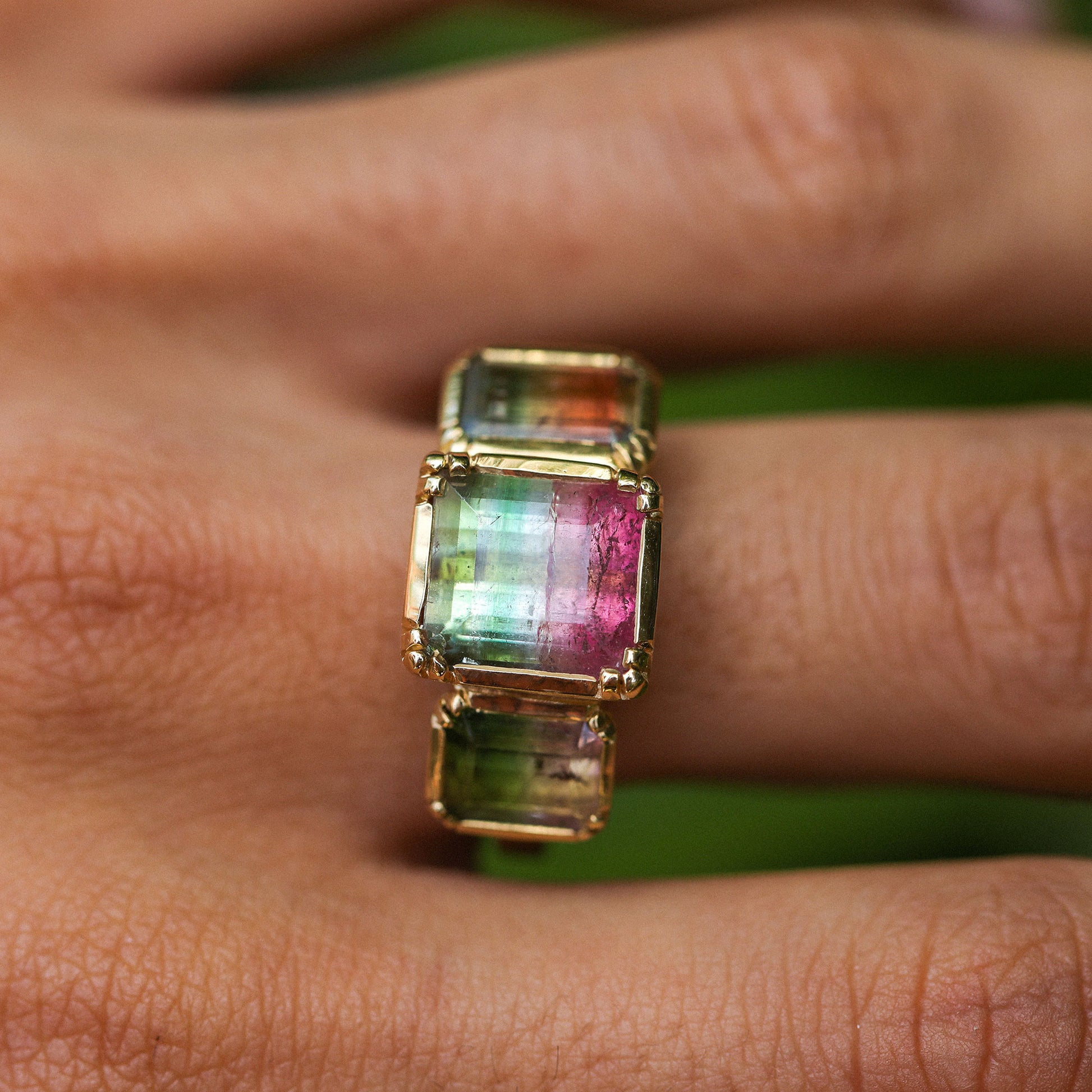 A gold ring with three rectangular iridescent bicolour tourmaline gemstones in green, pink, and blue tones, shown worn on a person's finger.