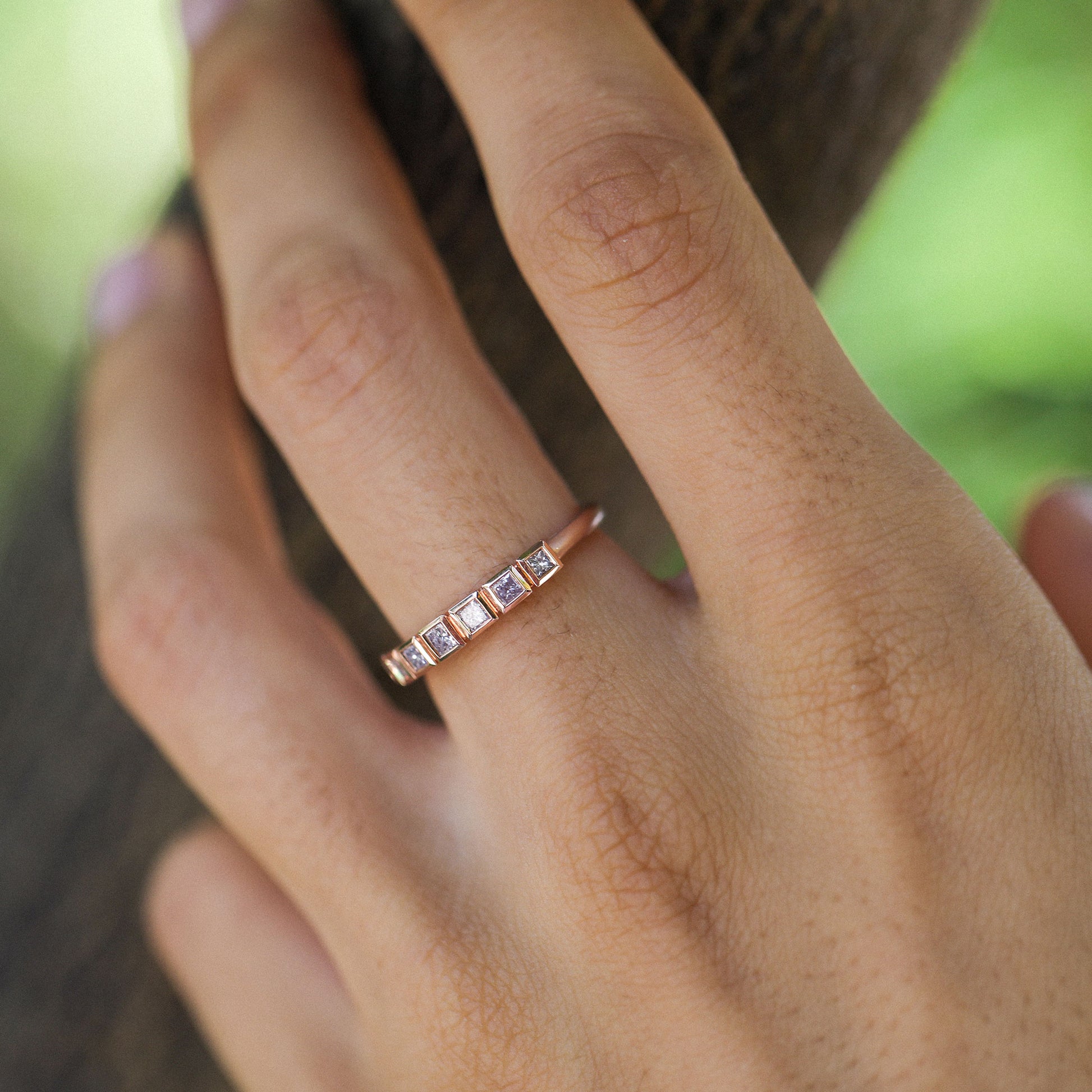 A close-up of a hand wearing a delicate rose gold band ring with five small square-cut pink diamond stones set in a row.