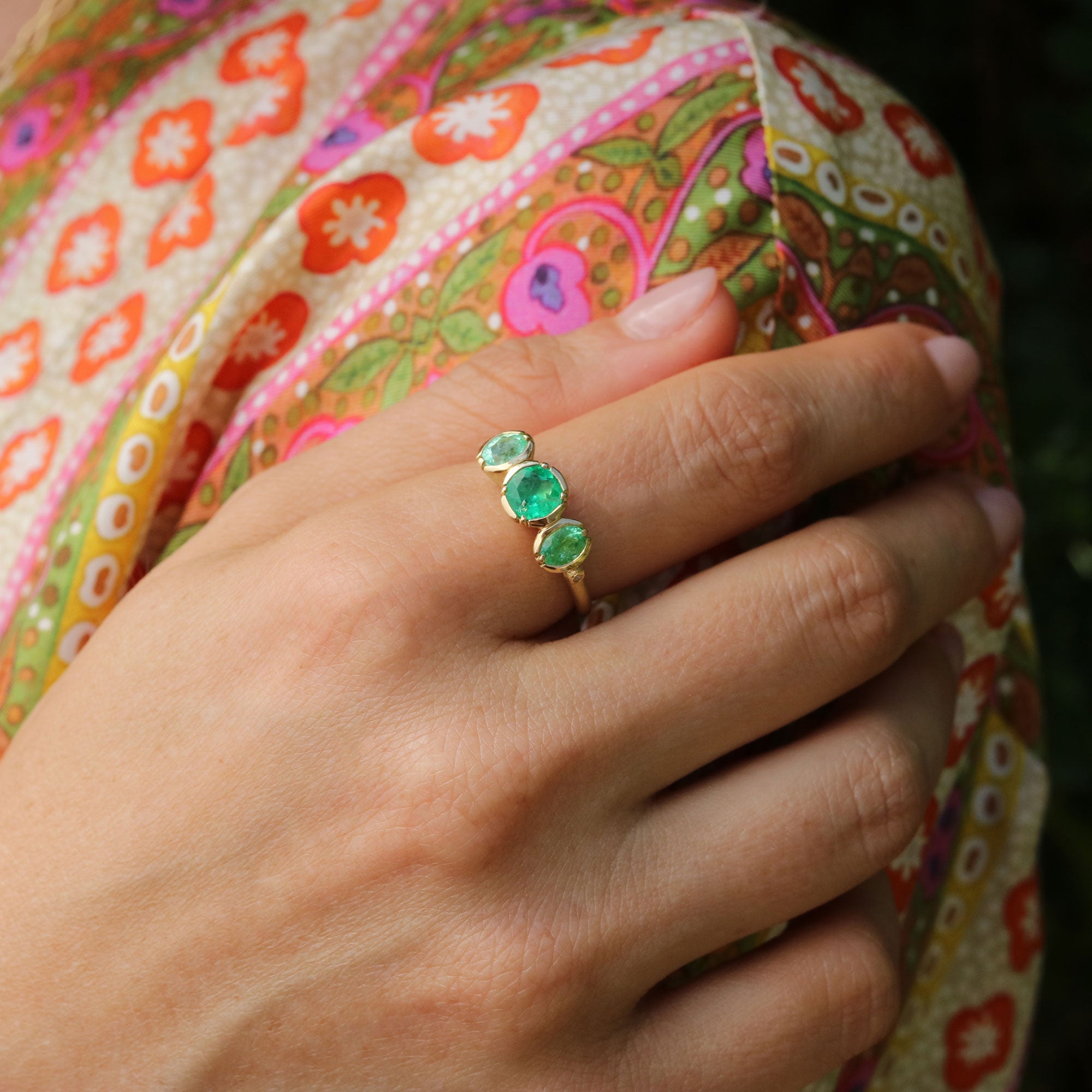 Hand wearing a ring with three green emerald and paraiba gemstones against a colorful fabric background