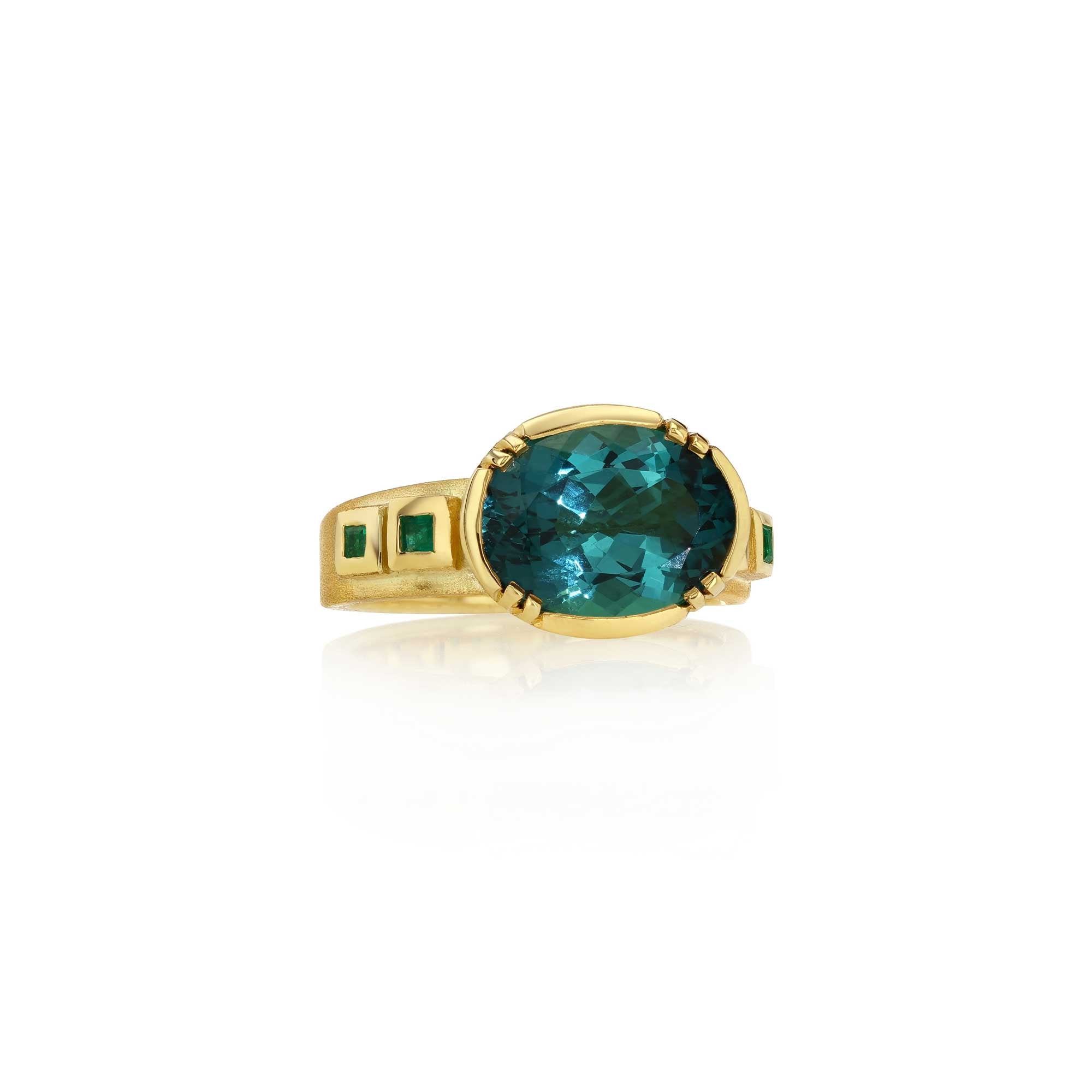 A yellow gold ring featuring a large oval-cut tourmaline gemstone in the center, with smaller square-cut emerald accents on the shoulders, set against a clean white background with a soft reflection.